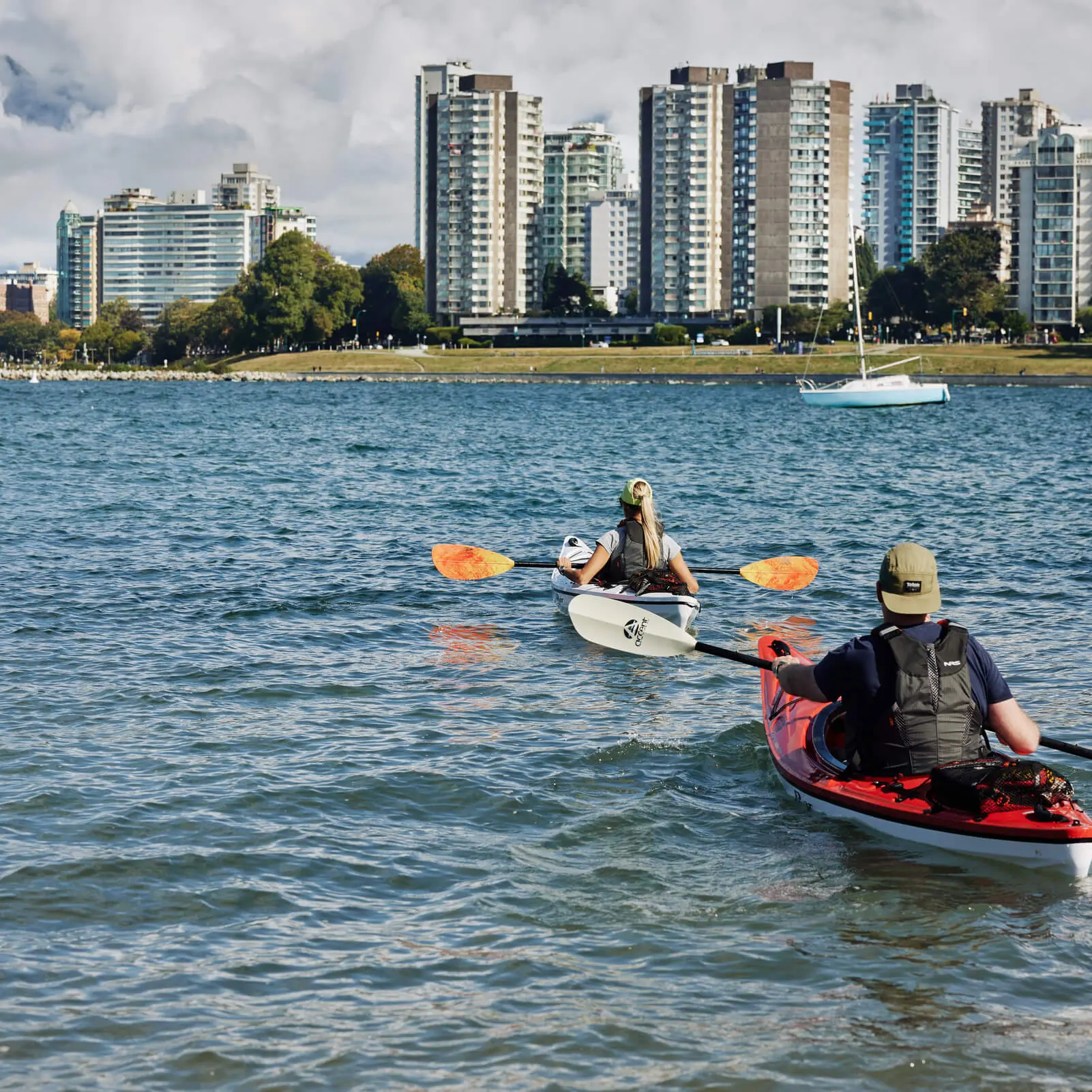 Two people kayaking on the water head toward a city shoreline with tall buildings and trees—a perfect moment to add to any Vancouver itinerary. A sailboat is anchored nearby under a partly cloudy sky.