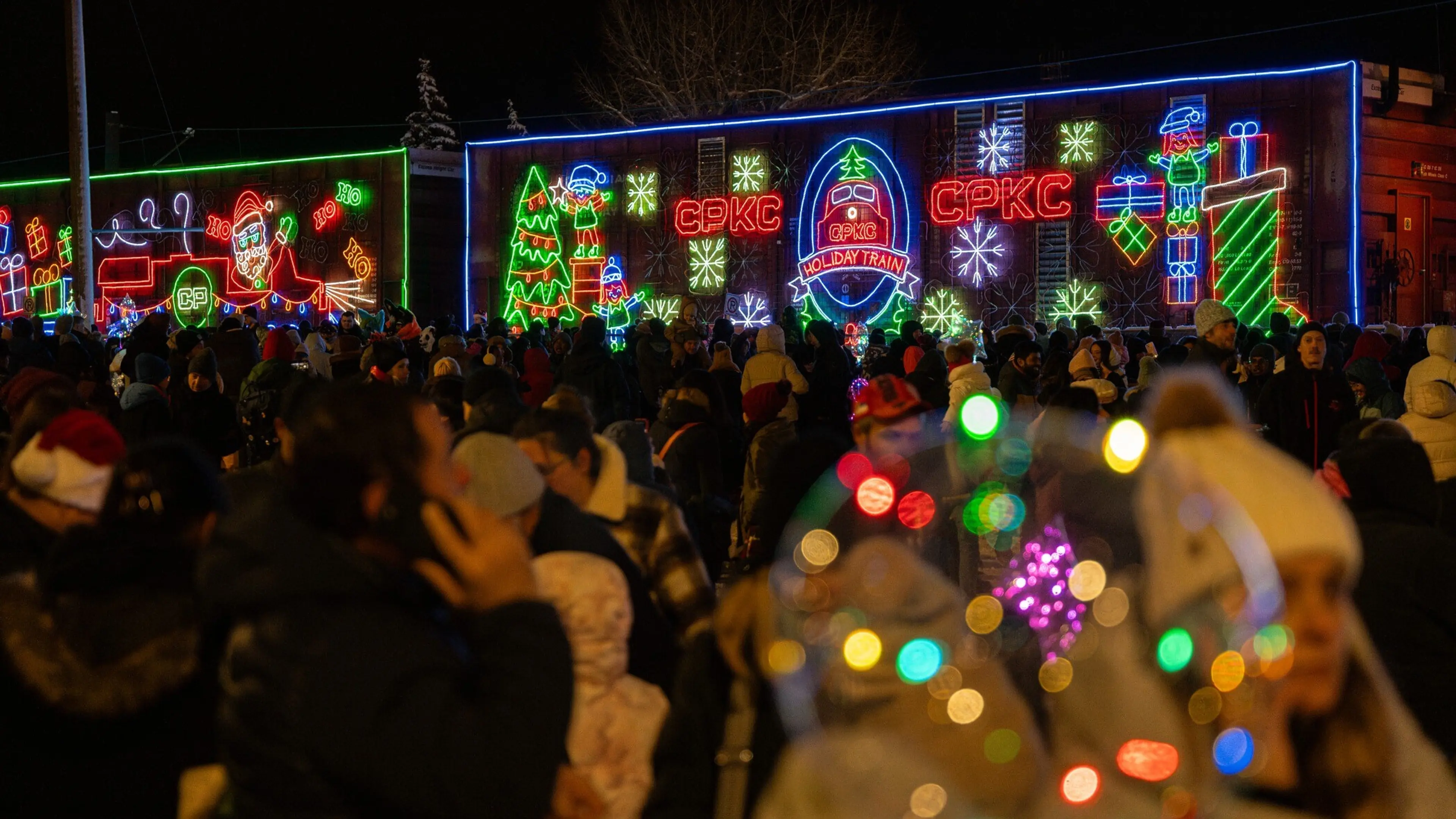 Crowd of people gathered at night in front of a train decorated with colorful holiday lights and festive designs.