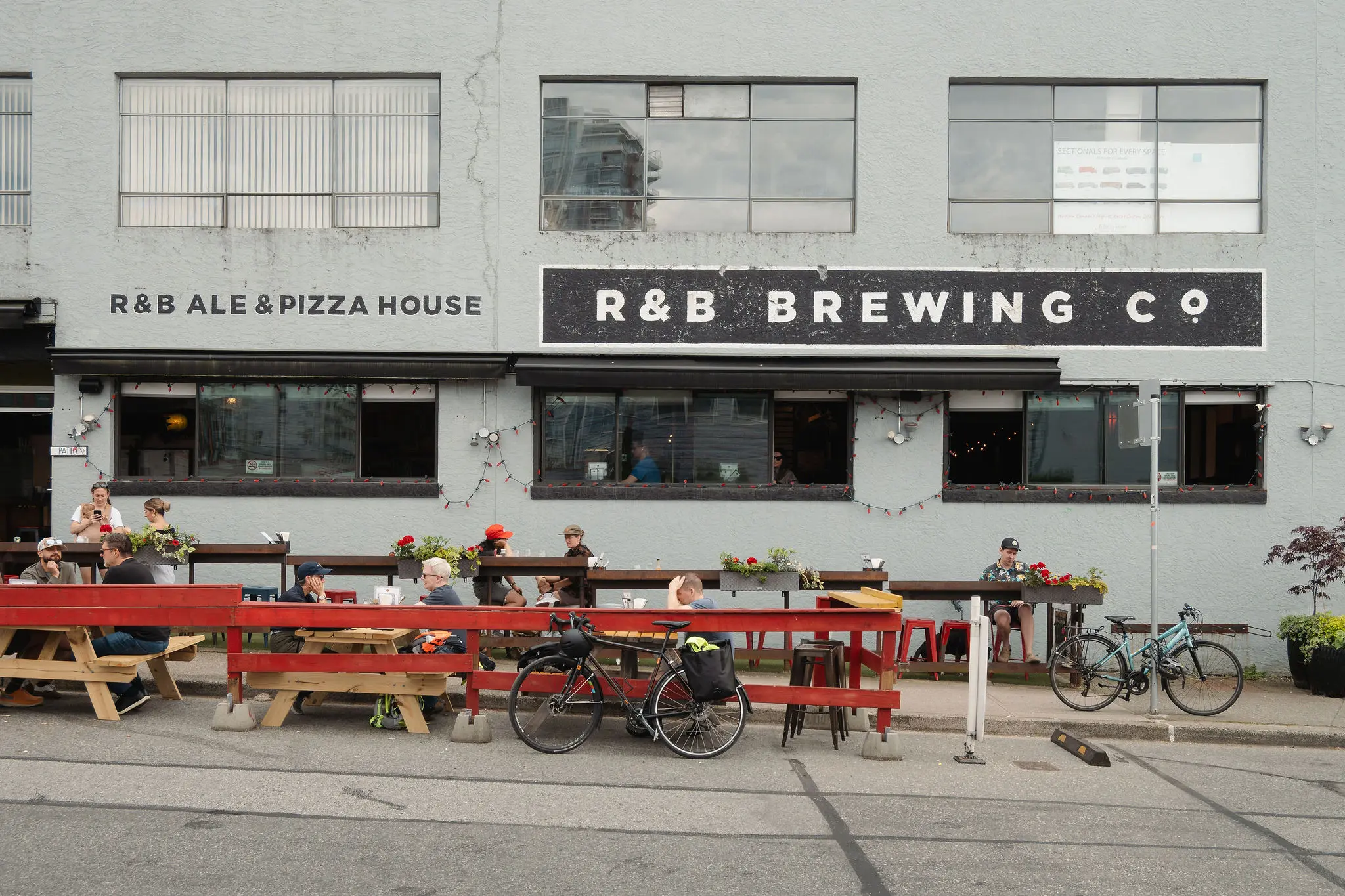 Outdoor seating area with people at wooden picnic tables in front of R&B Brewing Co building with bicycles parked nearby.