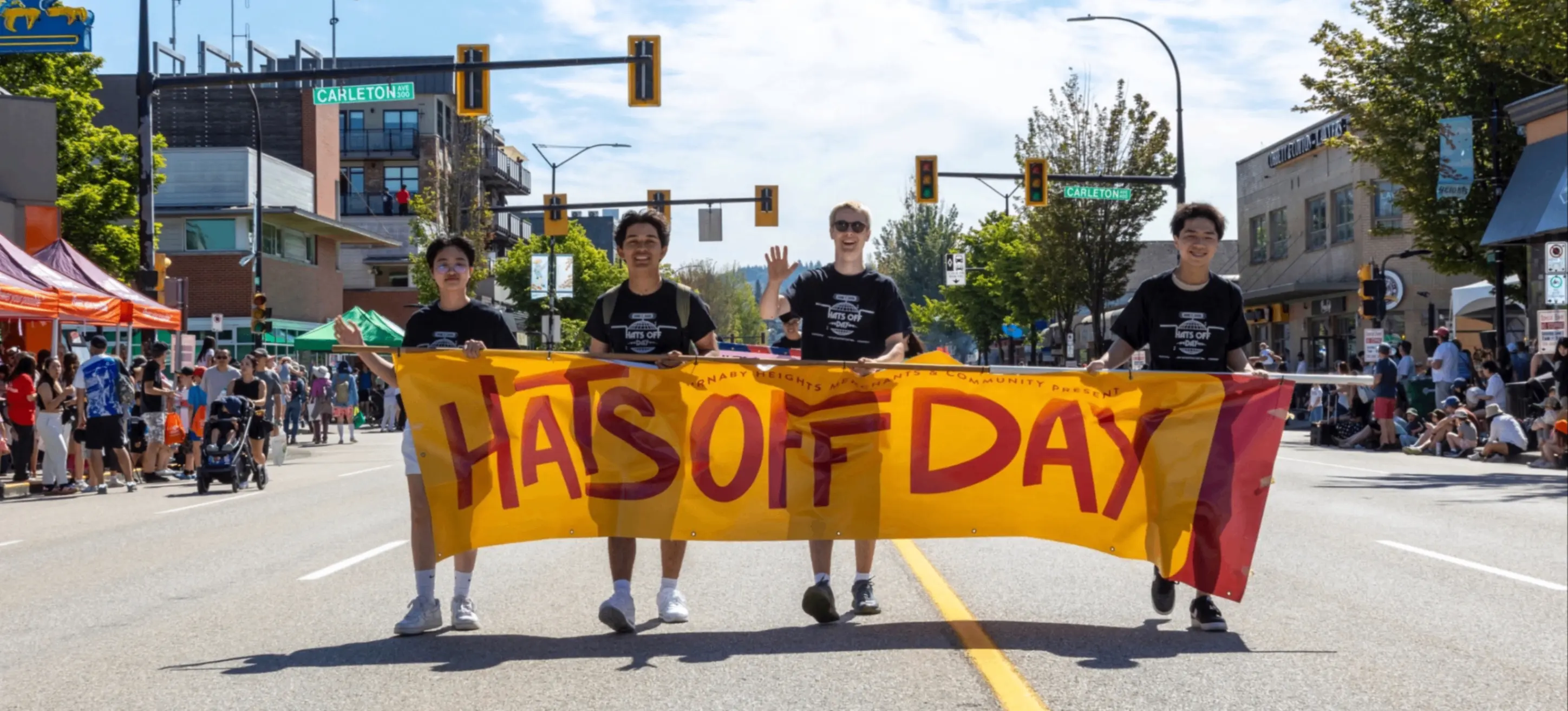 Four people walking on a street holding a yellow banner that reads "Hats Off Day" during a sunny outdoor event with crowds on both sides.