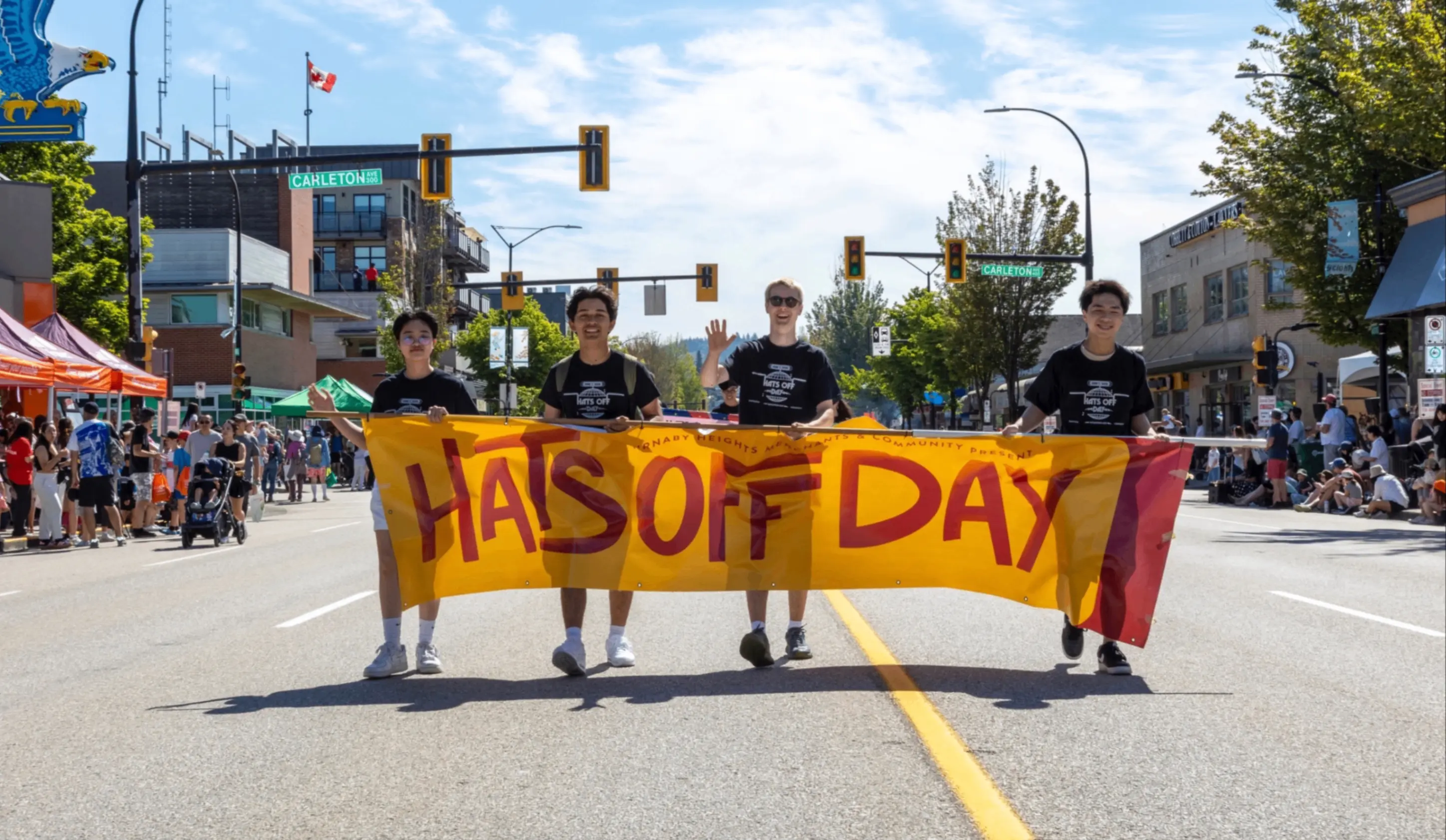 Four people walking on a street holding a yellow banner that reads "Hats Off Day" during a sunny outdoor event with crowds on both sides.