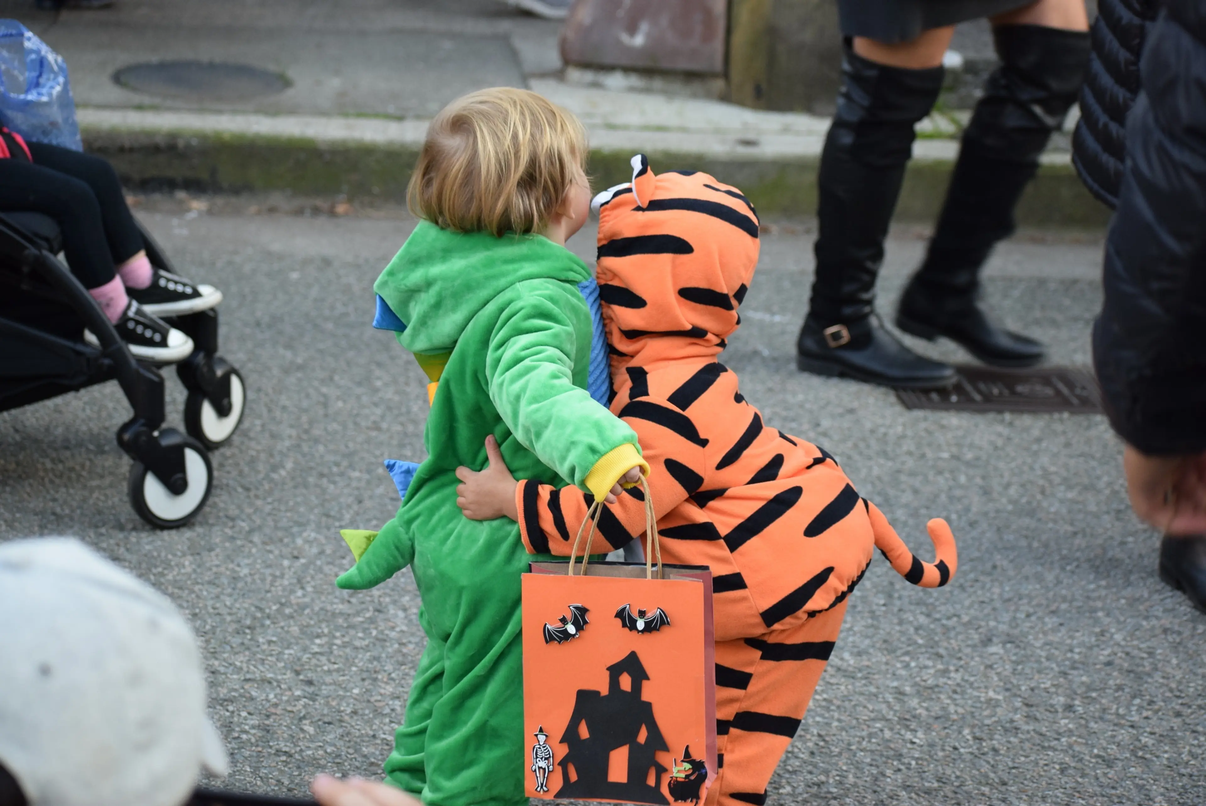 Two children in Halloween costumes, one in a green dinosaur suit and the other in an orange tiger suit, hugging on a street with a Halloween-themed bag.