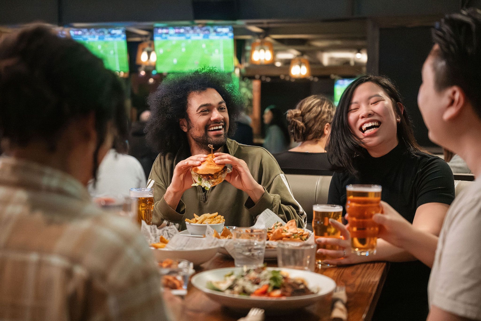 Four friends sitting at a table in a restaurant, one holding a burger, others holding glasses of beer, laughing and enjoying food.