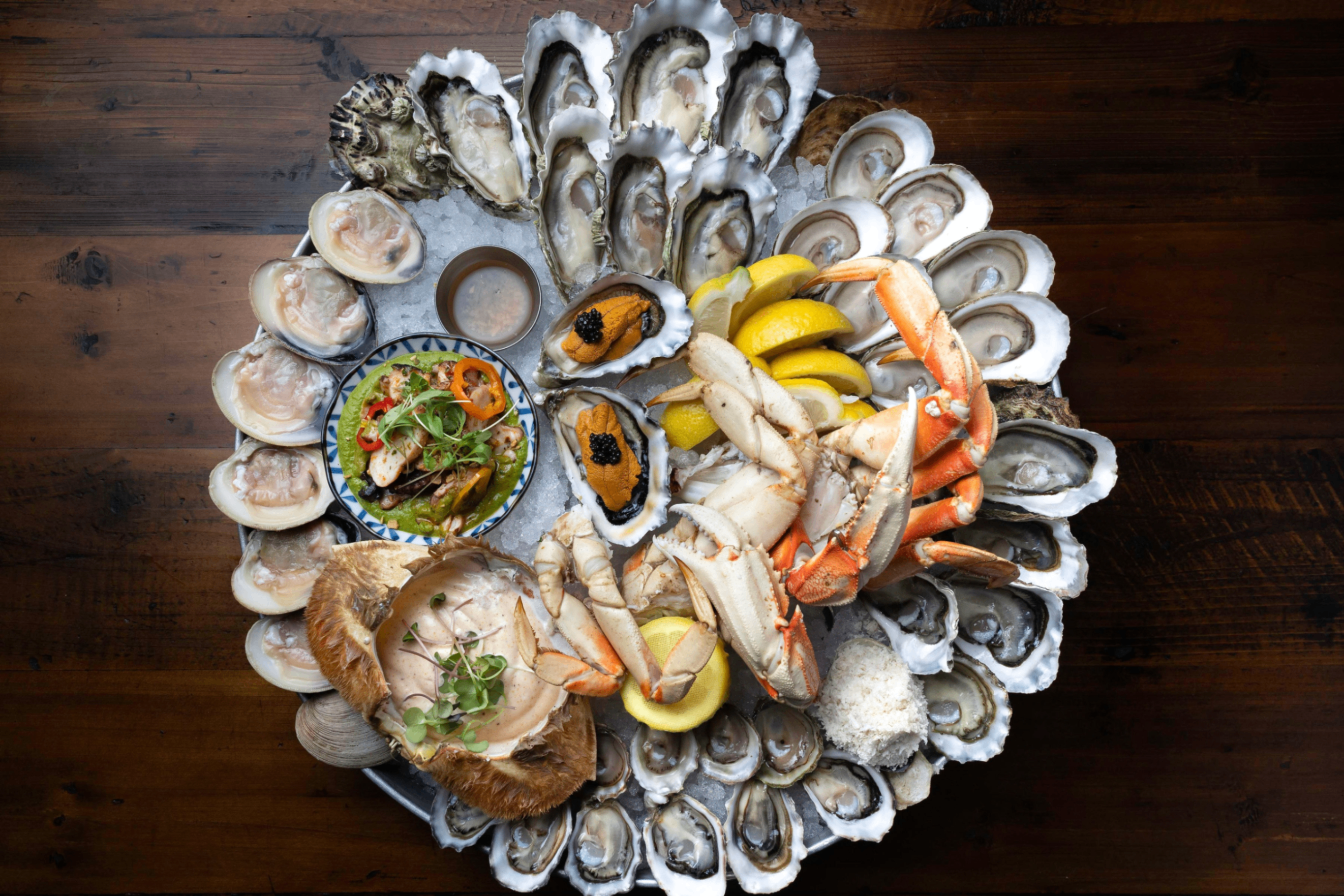 Seafood platter with crab legs, oysters, clams, lemon wedges, and a small bowl of sauce on a wooden table.