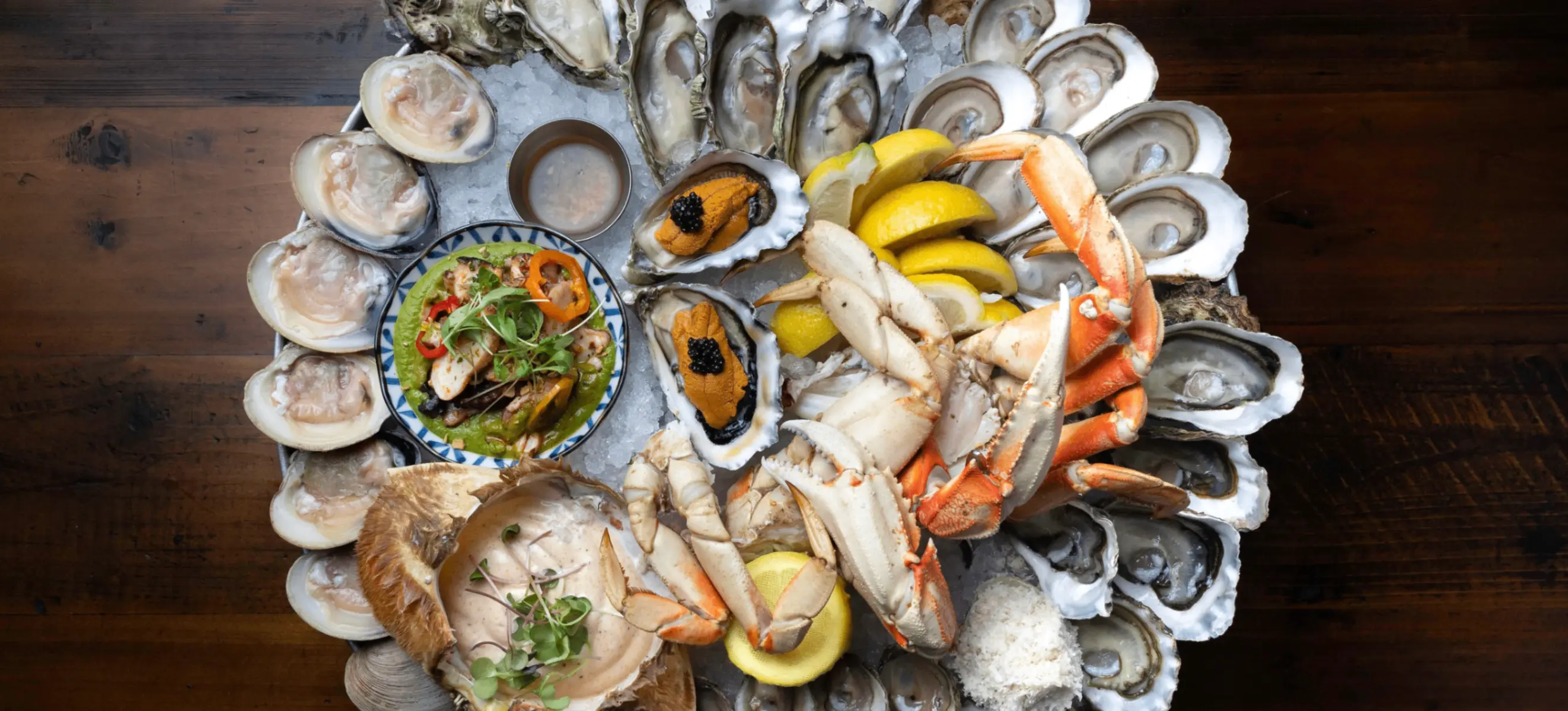Seafood platter with crab legs, oysters, clams, lemon wedges, and a small bowl of sauce on a wooden table.