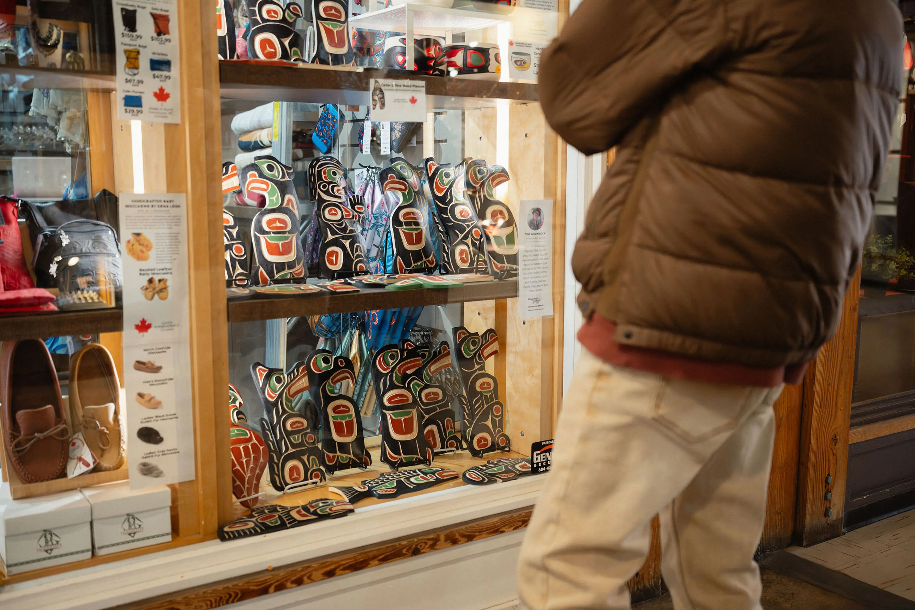 Person in brown jacket and white pants looking at a display of Indigenous art pieces in a store window.