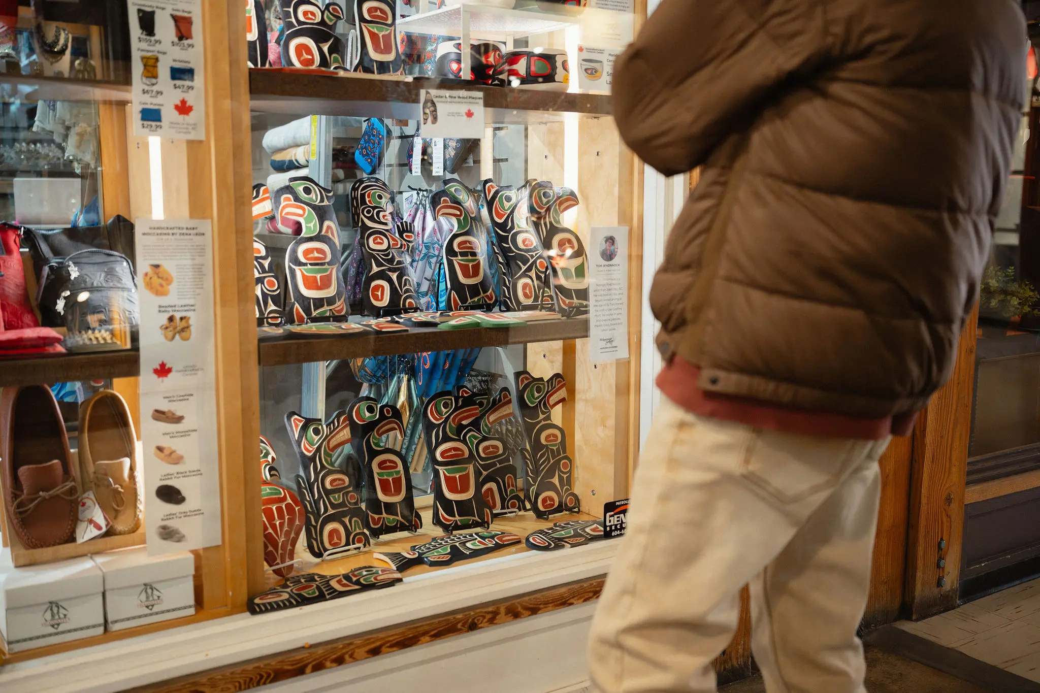 Person in brown jacket and white pants looking at a display of Indigenous art pieces in a store window.