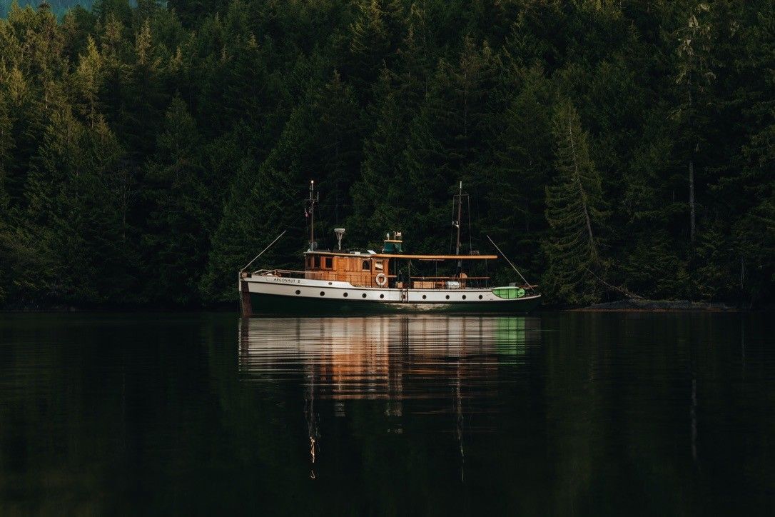 A wooden boat named Argonaut II anchored on calm water with dense evergreen forest in the background.