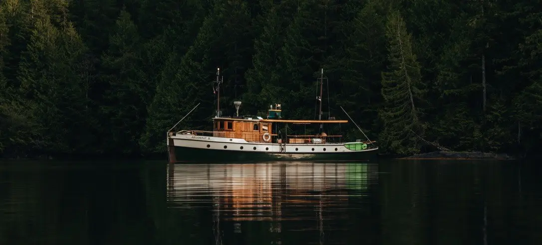 A wooden boat named Argonaut II anchored on calm water with dense evergreen forest in the background.