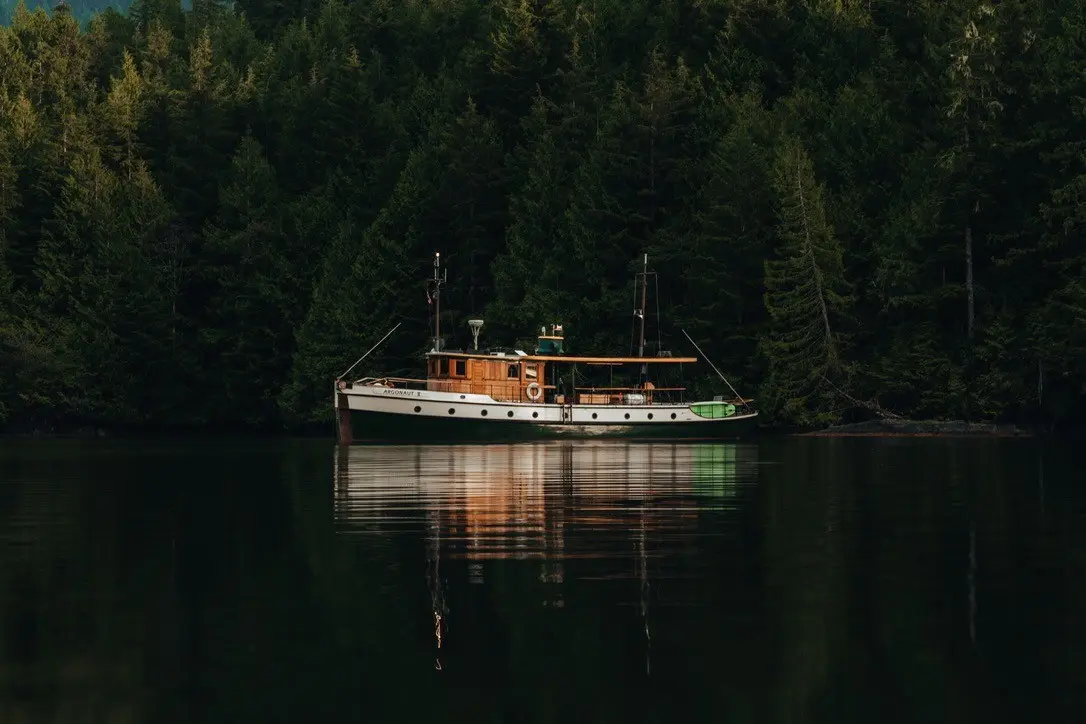 A wooden boat named Argonaut II anchored on calm water with dense evergreen forest in the background.