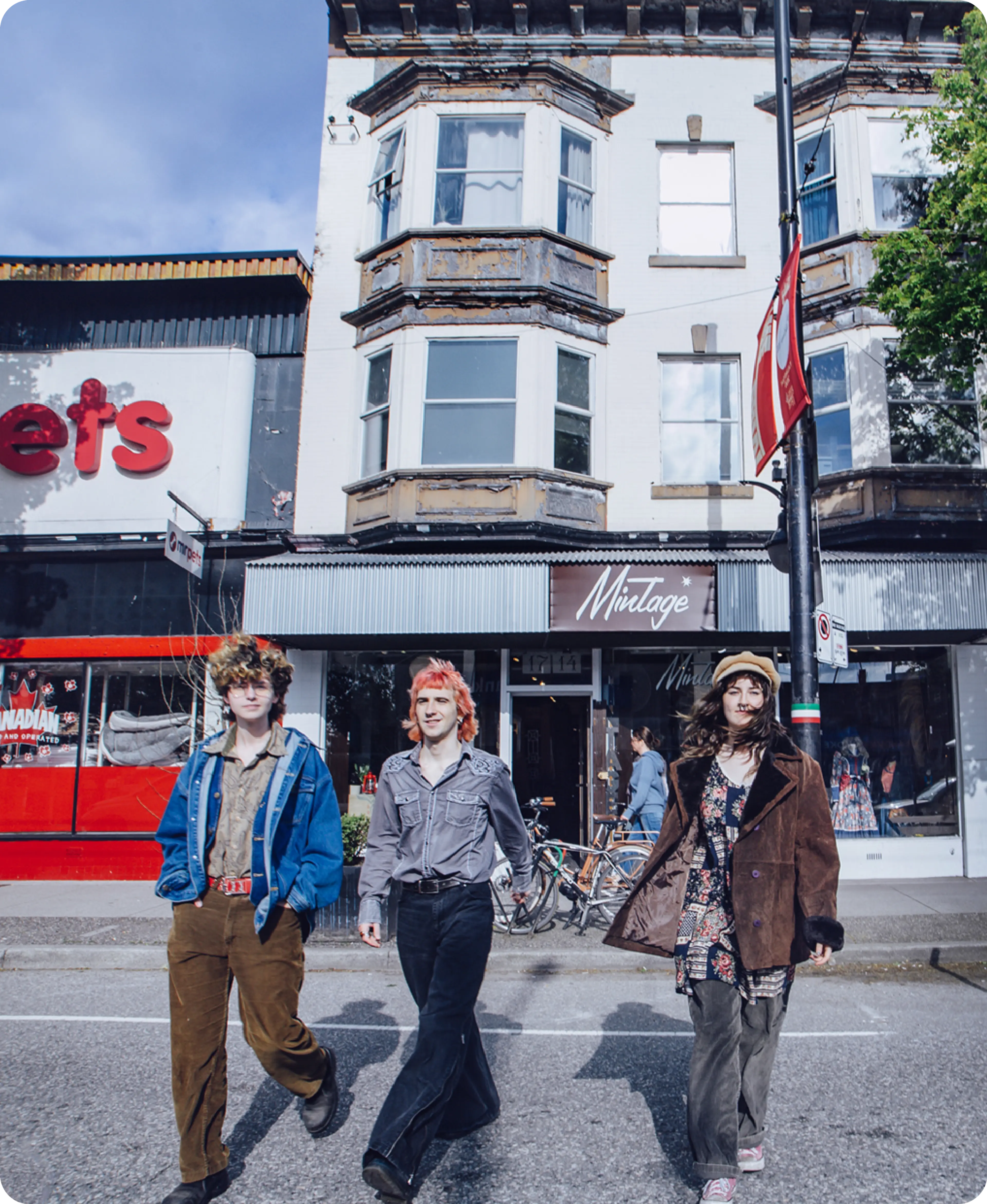 Three people walking on a street in front of a vintage store with bay windows above and a red storefront to the left.