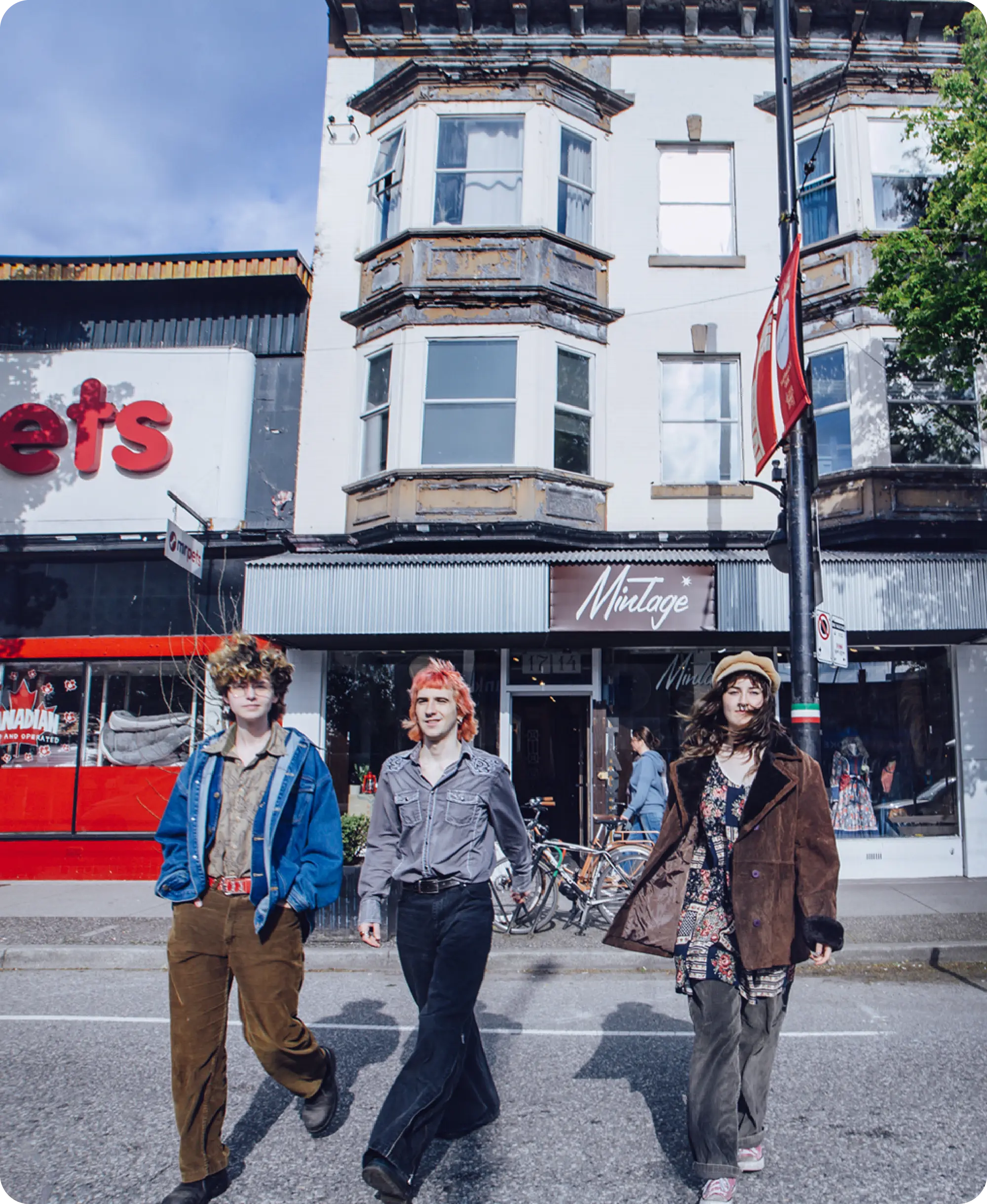 Three people walking on a street in front of a vintage store with bay windows above and a red storefront to the left.