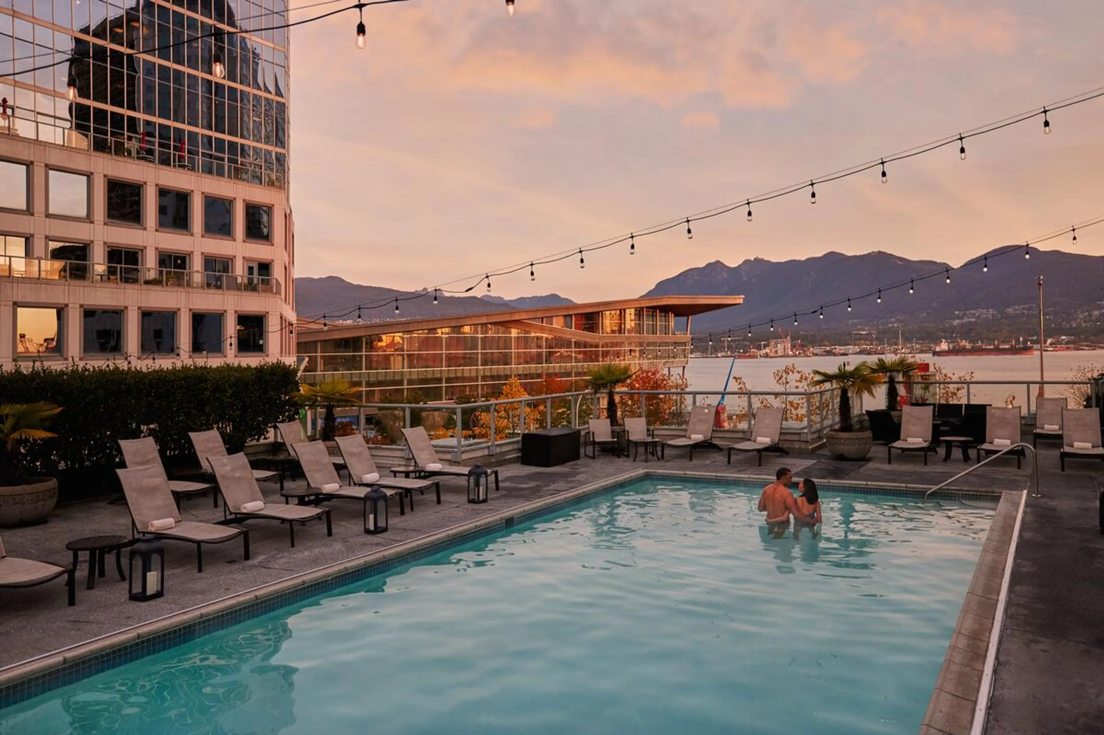 Two people standing in a rooftop pool at sunset with lounge chairs, string lights, and mountains in the background.