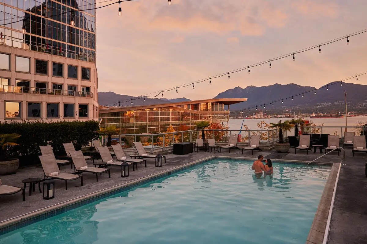 Two people standing in a rooftop pool at sunset with lounge chairs, string lights, and mountains in the background.