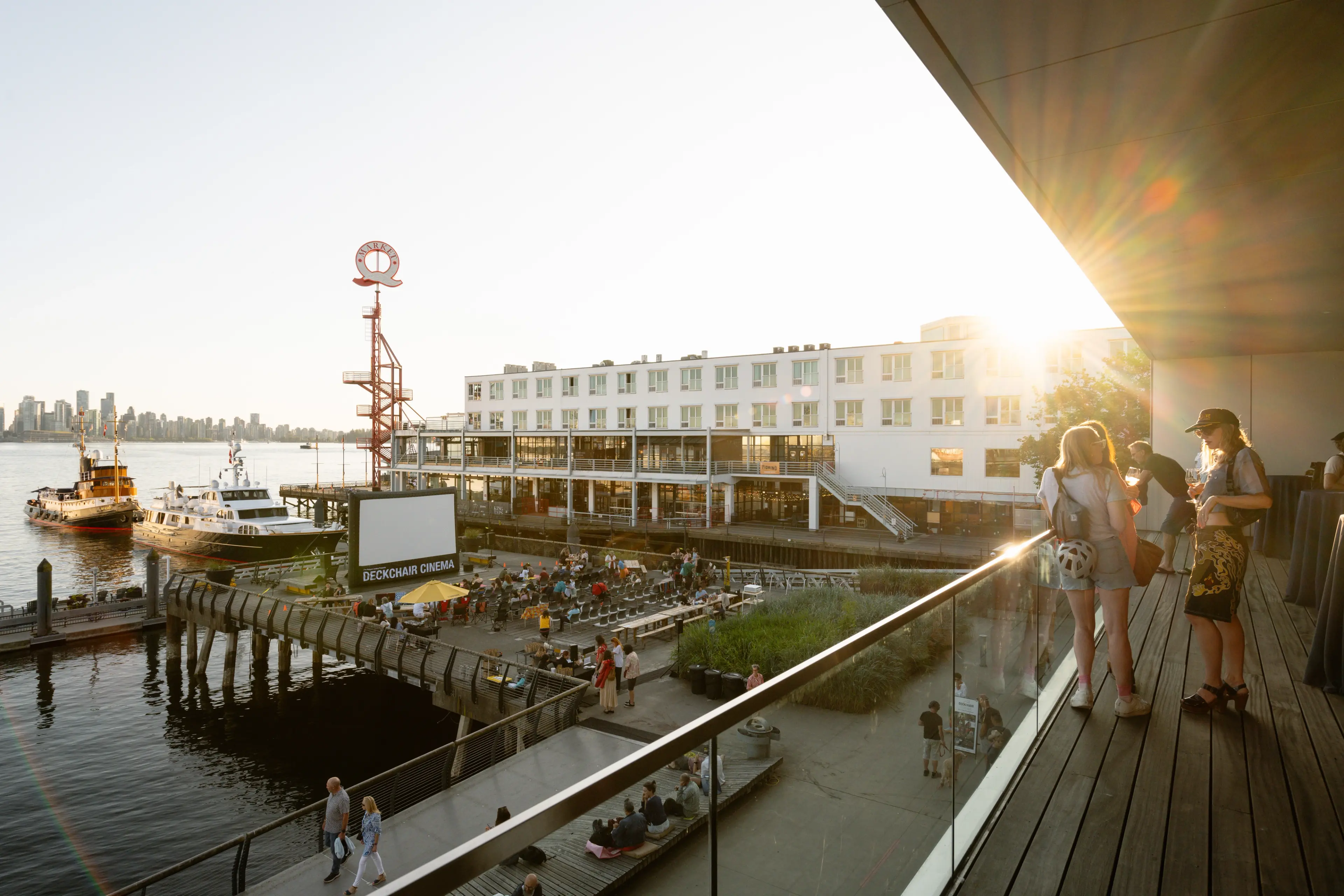 People gathered at an outdoor deckchair cinema by the water during sunset, with boats docked nearby and a city skyline in the background.