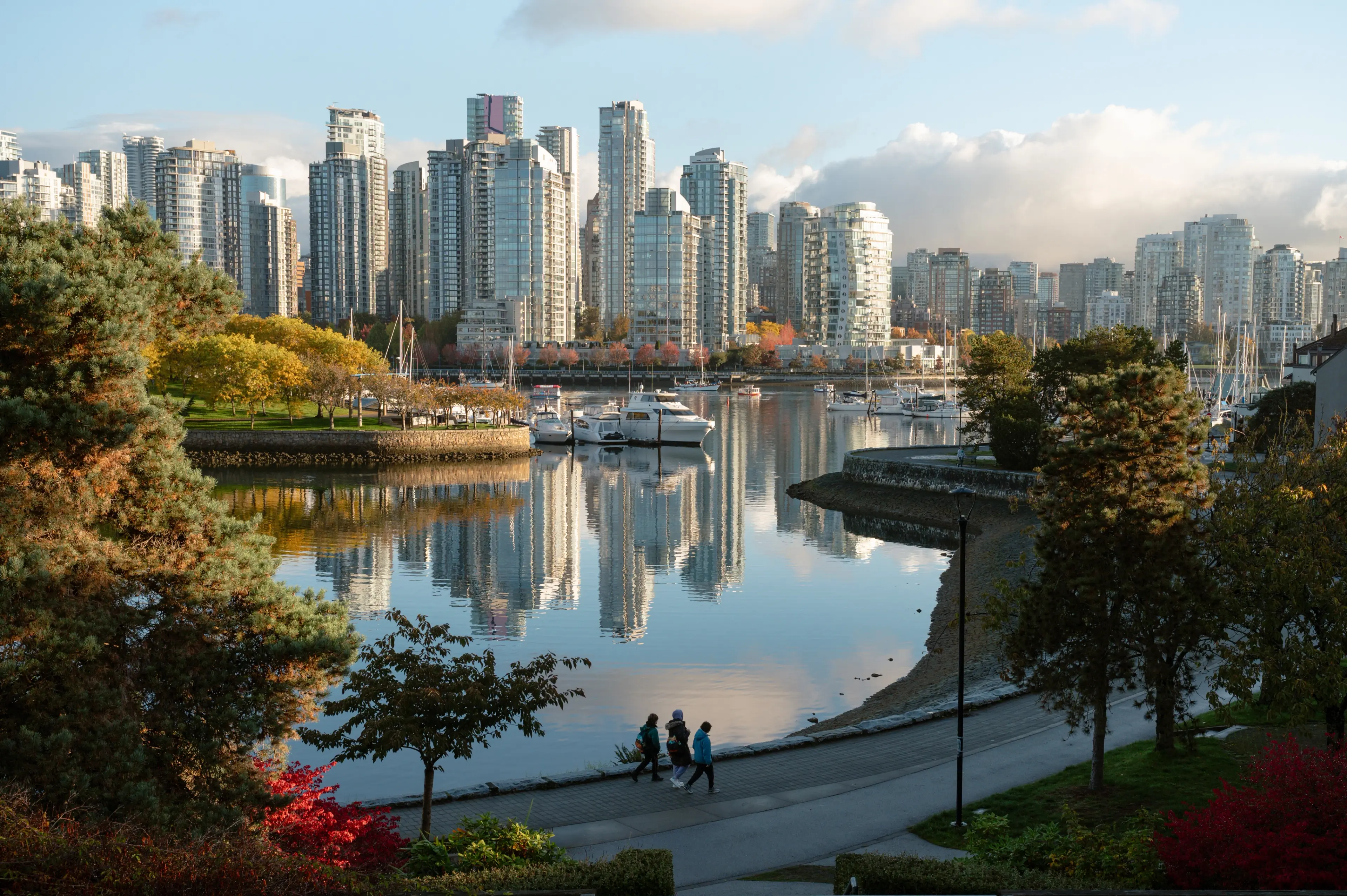 Three people walking on a path beside a calm water body with boats and high-rise buildings reflected in the water.