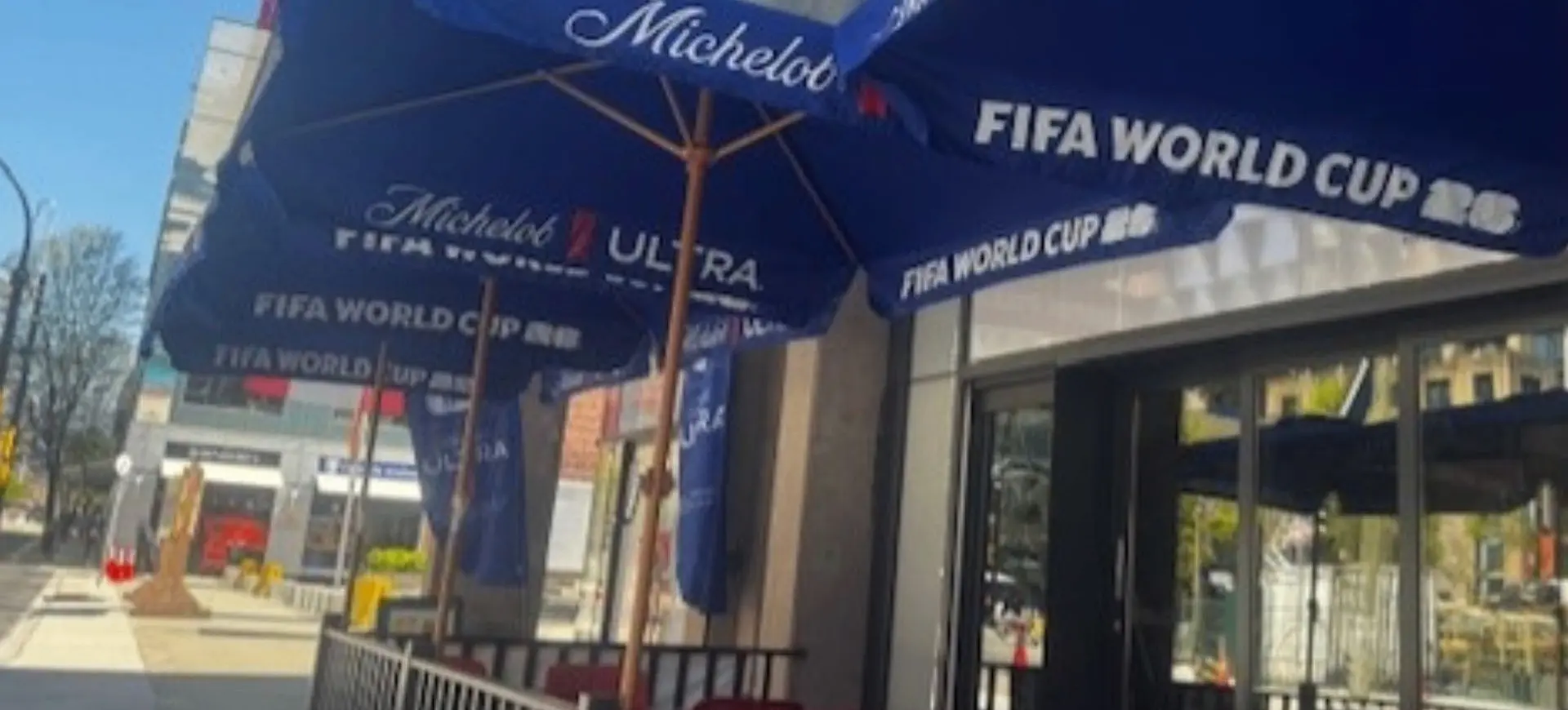 Outdoor seating area with blue umbrellas displaying Michelob Ultra and FIFA World Cup 2026 logos on a sunny day.