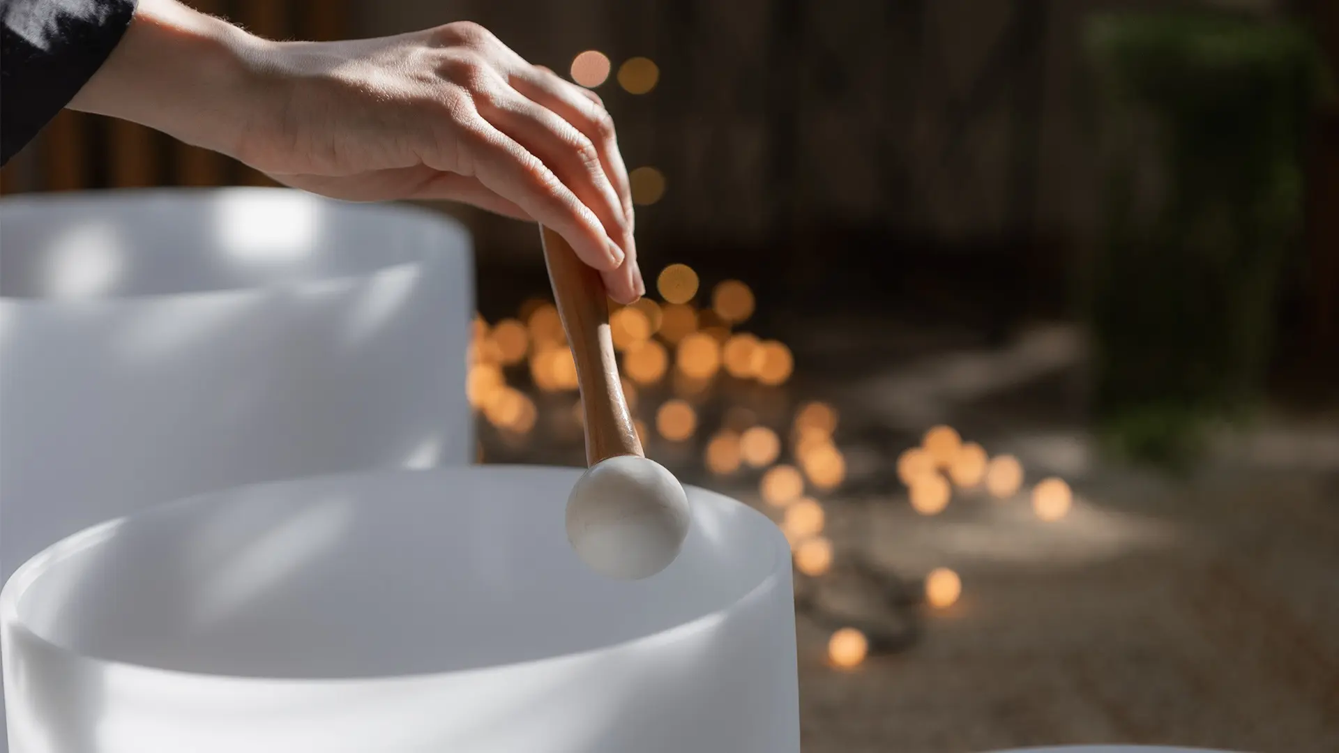 Hand holding a wooden mallet striking a white crystal singing bowl with blurred warm lights in the background.