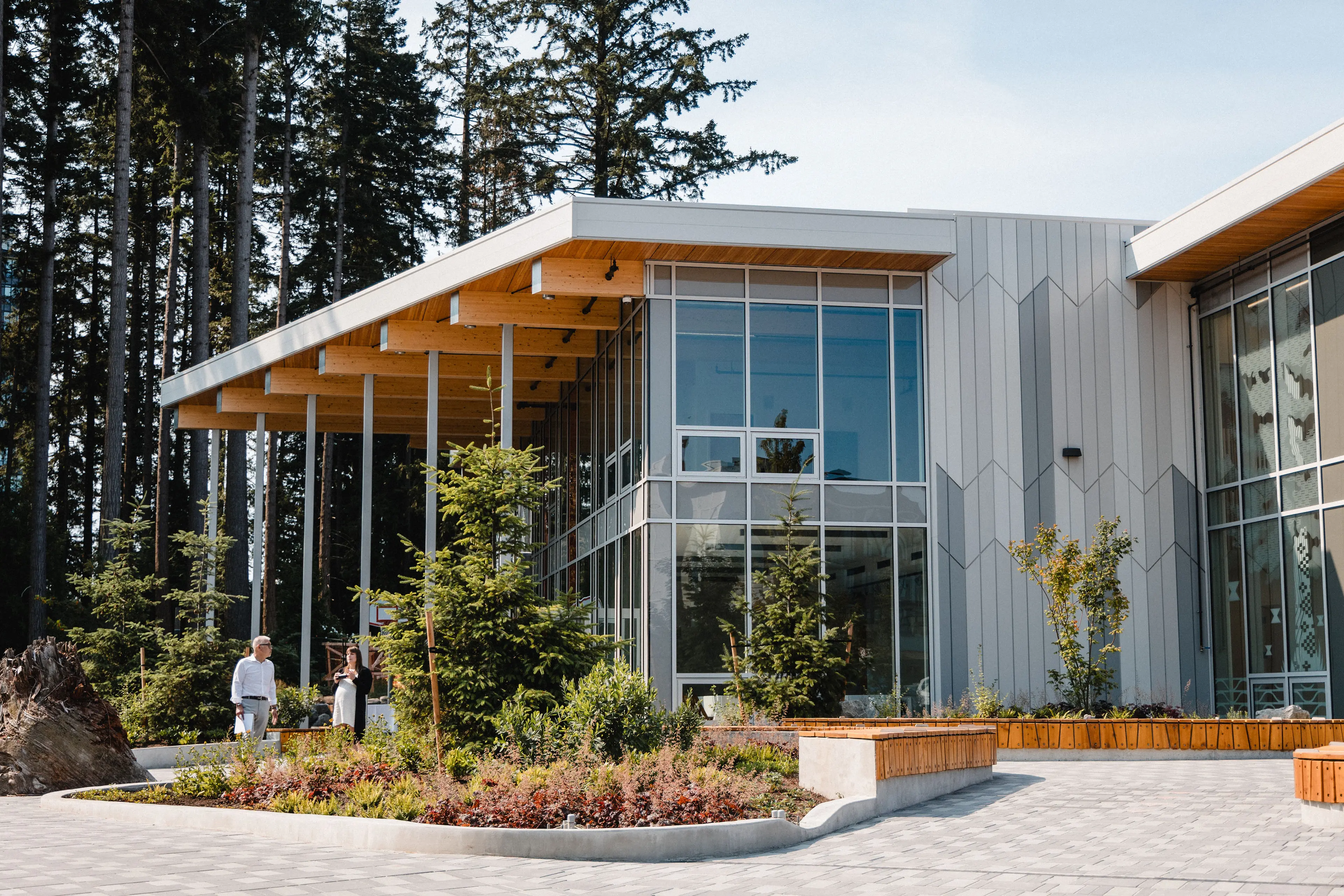 Modern building with large glass windows and wooden roof beams surrounded by trees and landscaped plants, two people walking nearby.