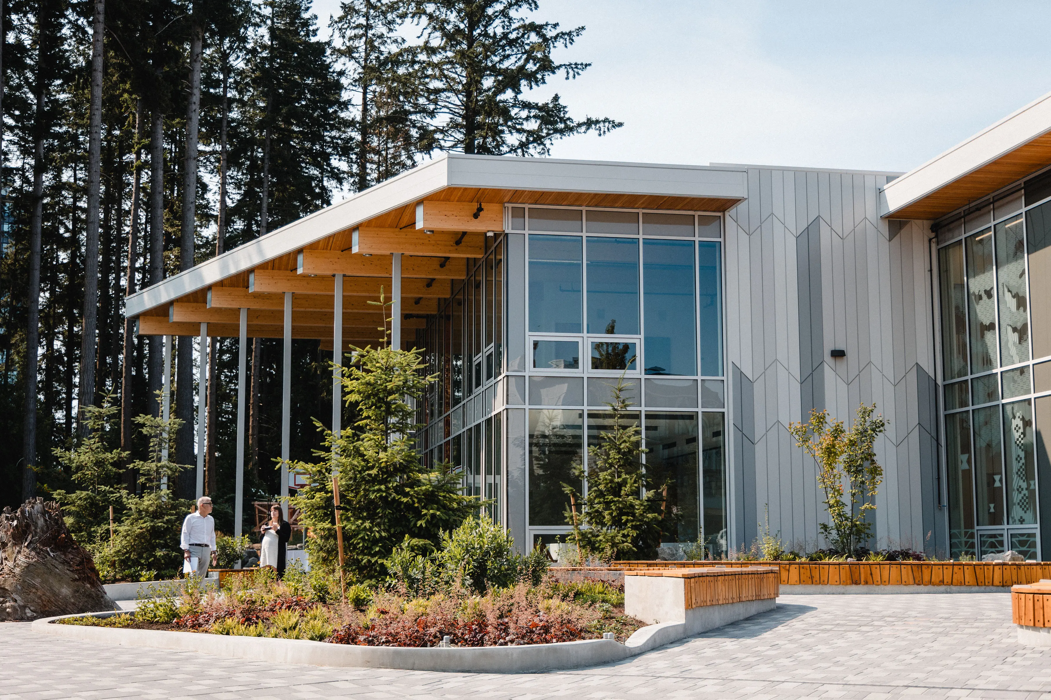 Modern building with large glass windows and wooden roof beams surrounded by trees and landscaped plants, two people walking nearby.