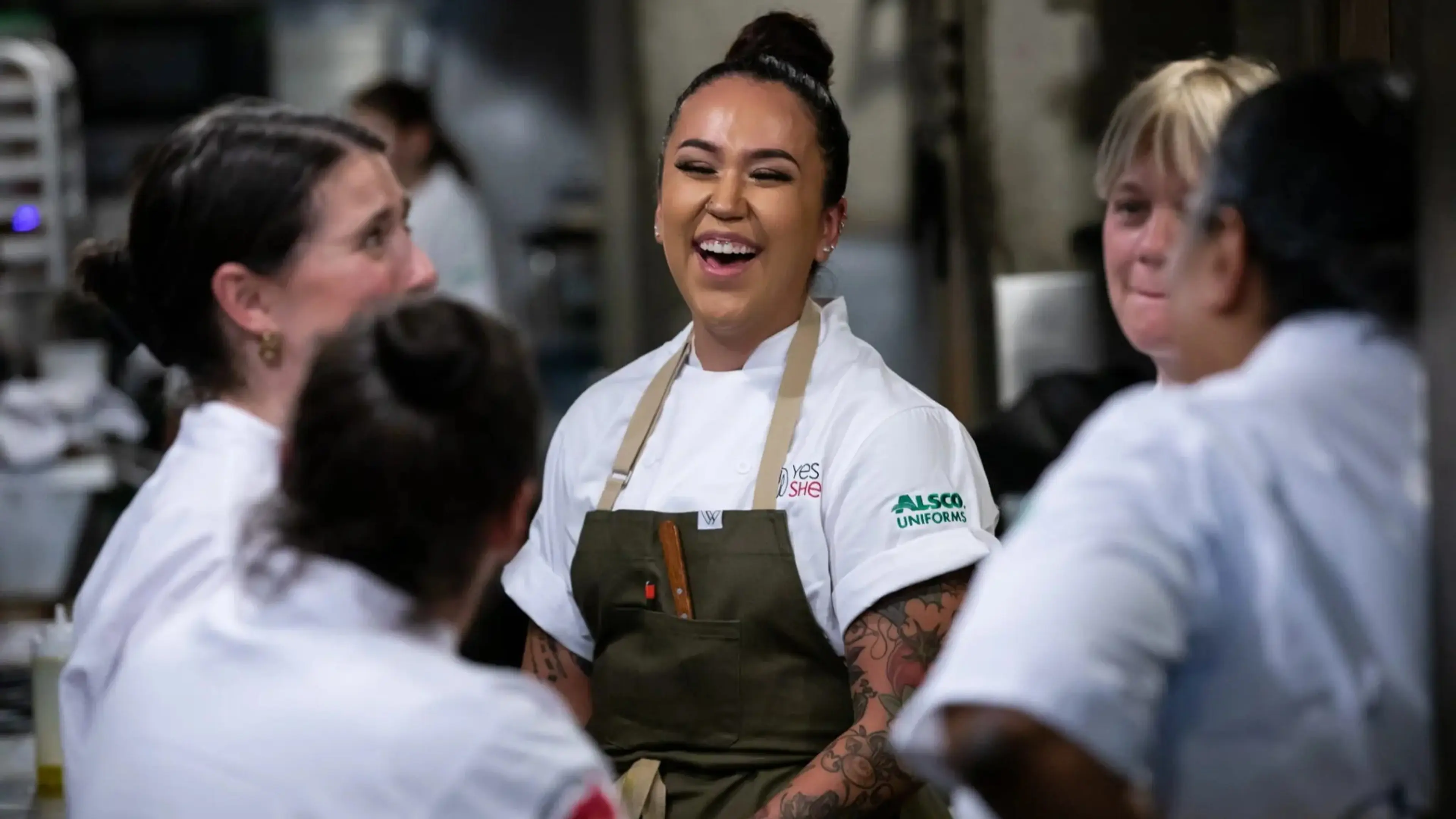 Five people in chef uniforms standing in a kitchen, one person in the center laughing with tattoos visible on arms.