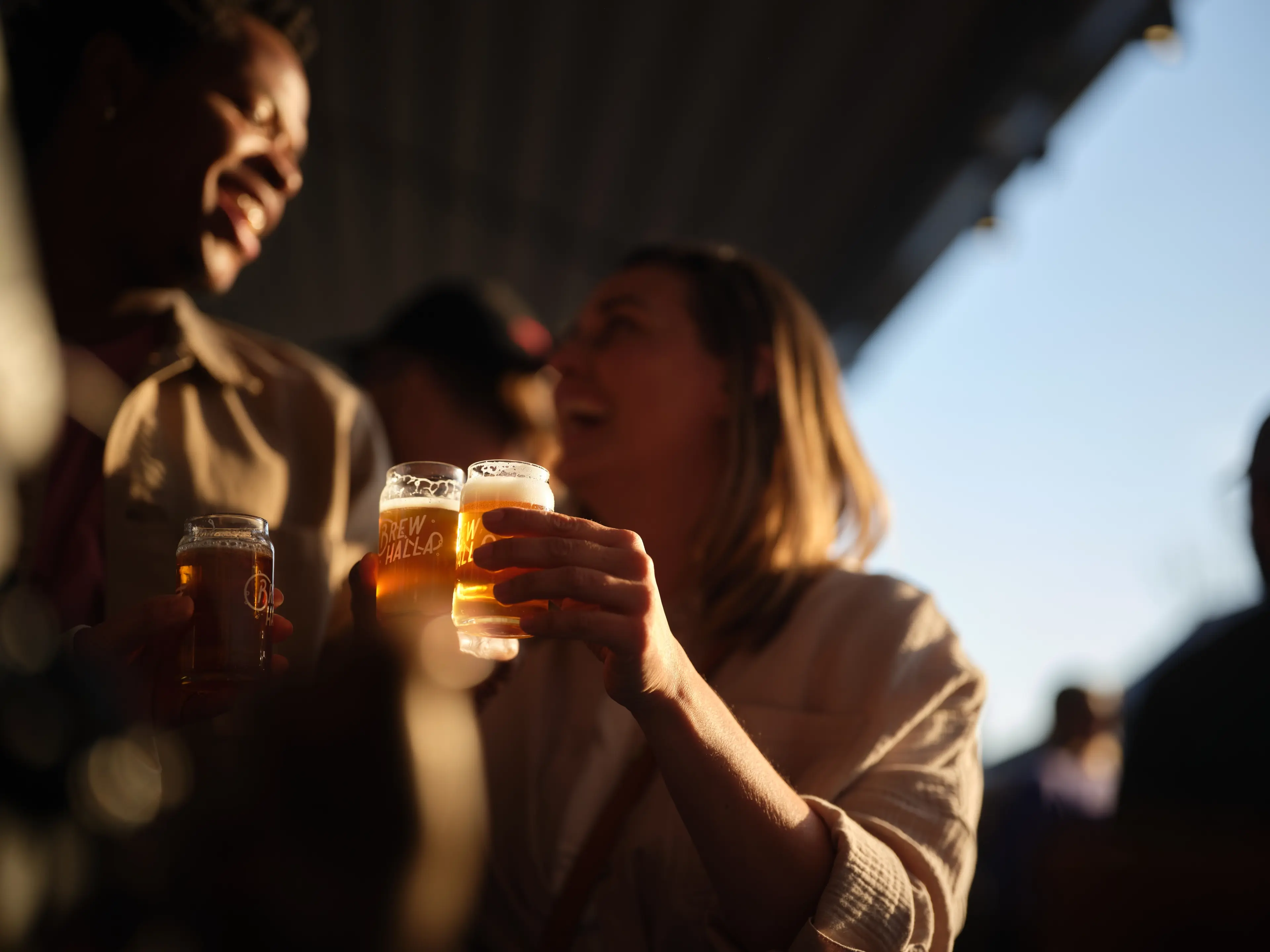 Two people holding glasses of beer and smiling in a dimly lit setting with a bright sky in the background.