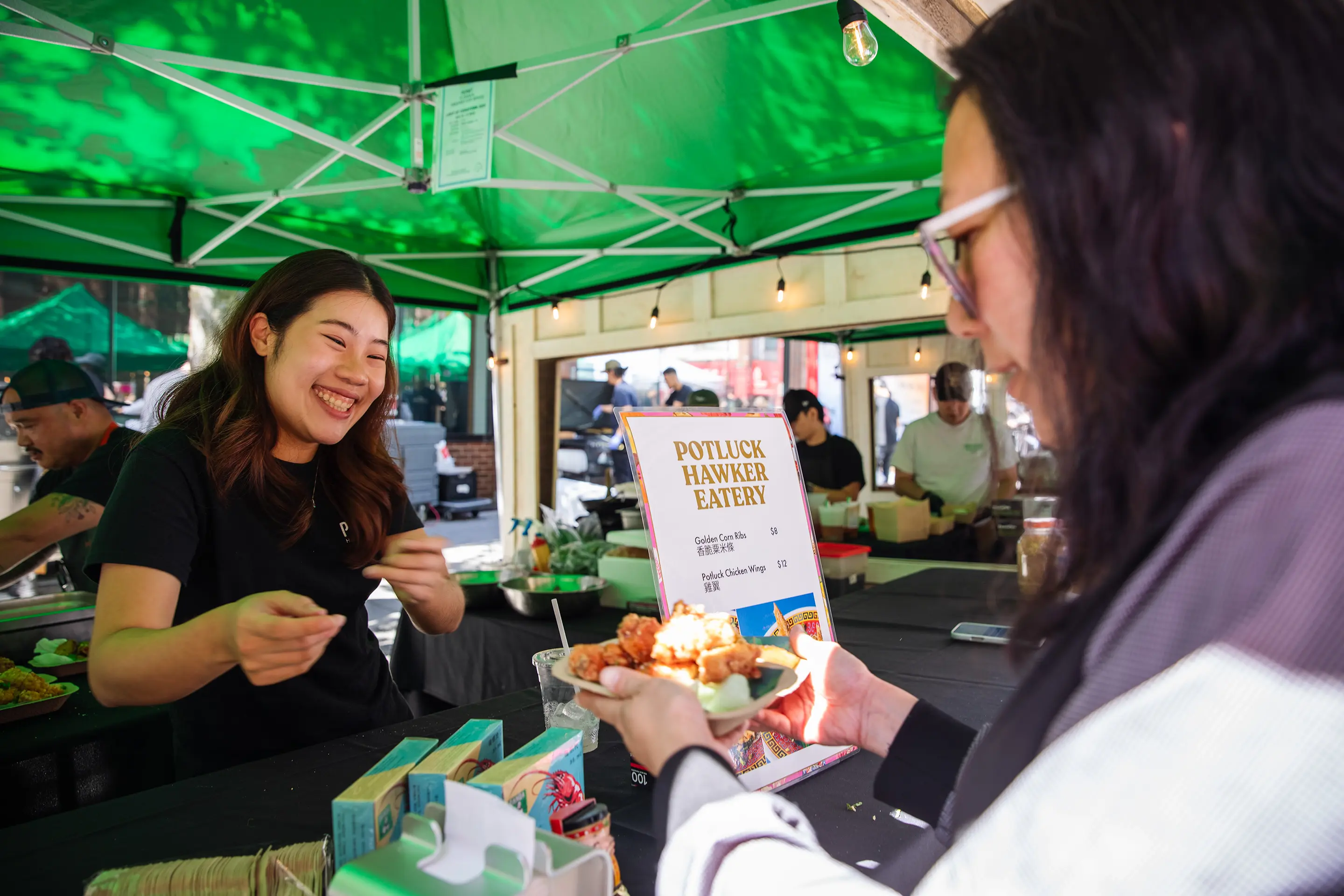 A person serving fried food to a smiling vendor under a green tent at Potluck Hawker Eatery food stall.