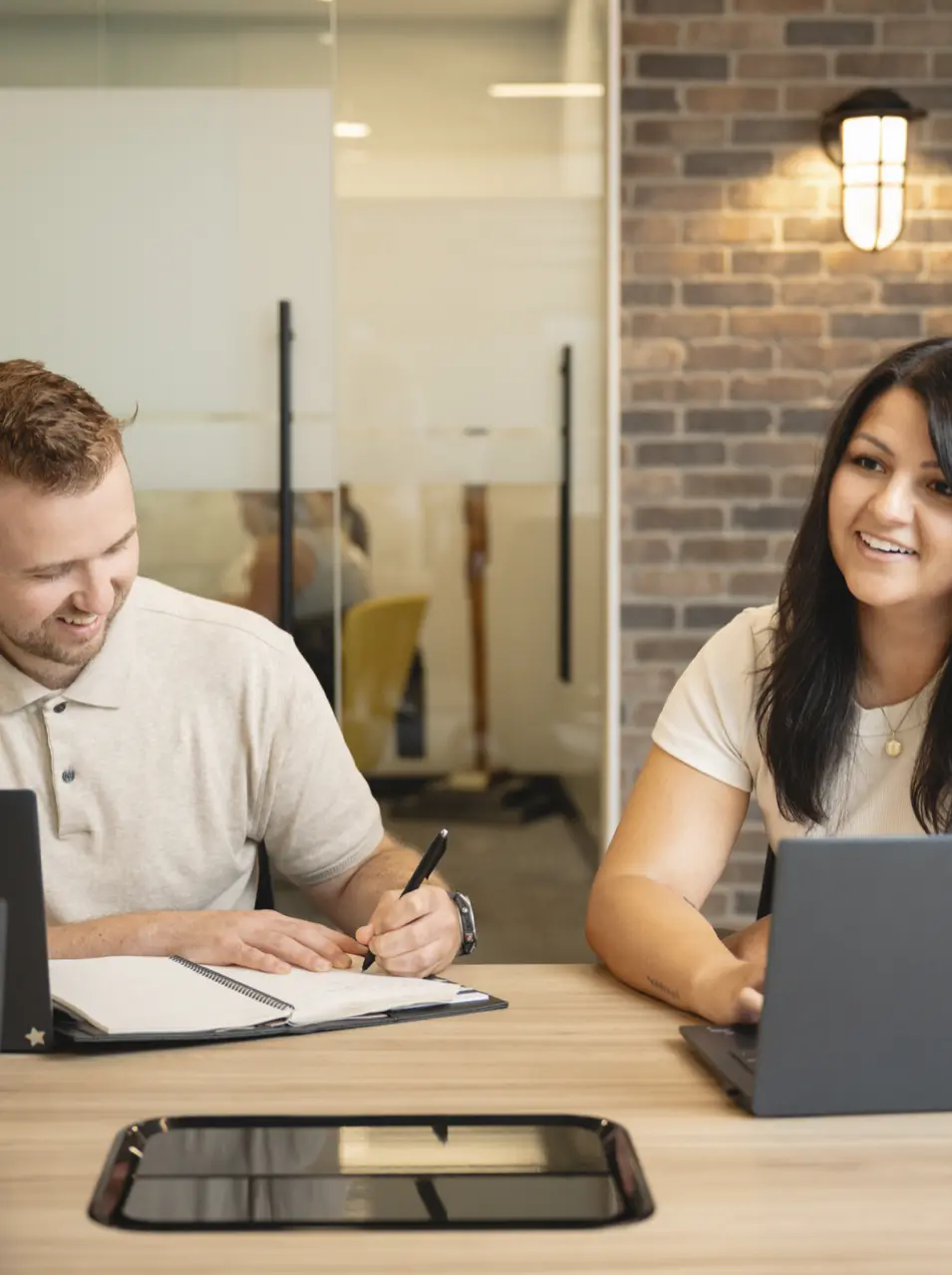 Two people sitting at a table with laptops and a notebook, one writing and the other smiling, in a modern office.