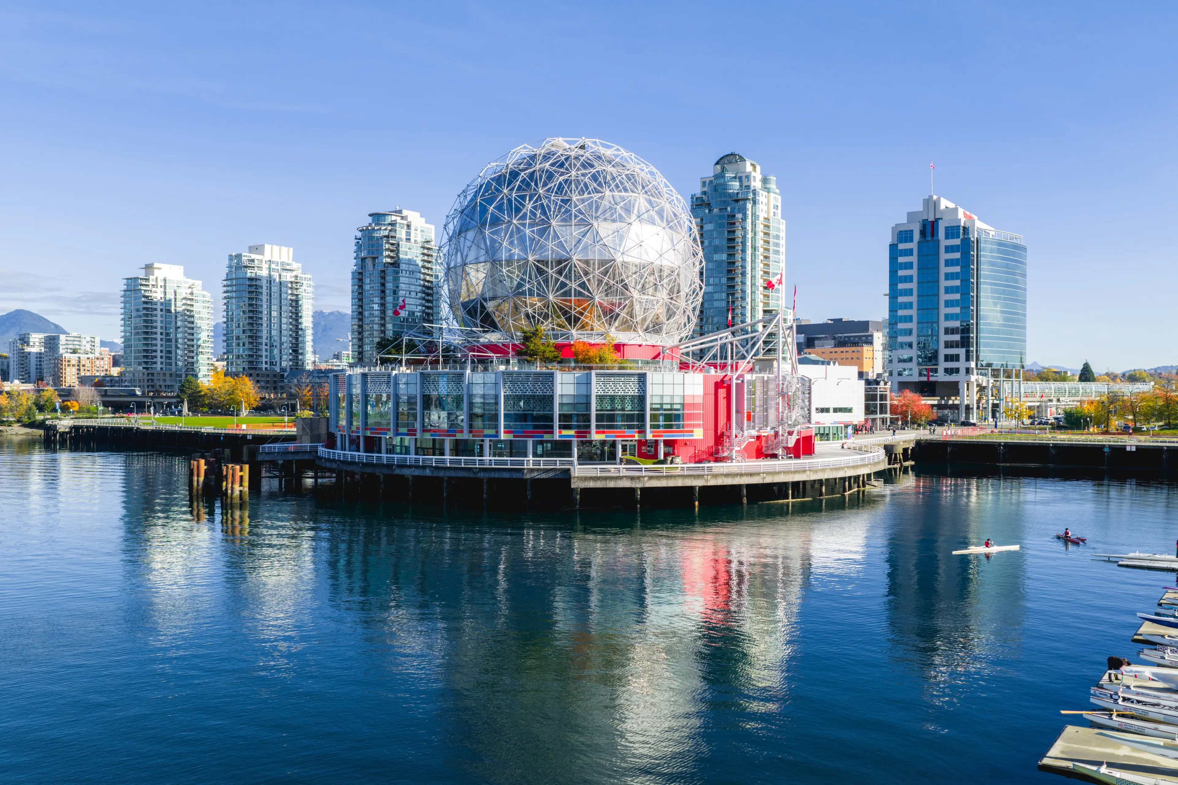 Geodesic dome building on waterfront with modern high-rise buildings and kayakers on calm water under clear sky.