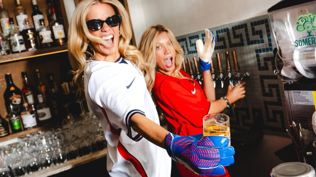 Two people in sports jerseys and gloves serving beer from taps behind a bar, one holding a glass of beer and smiling.