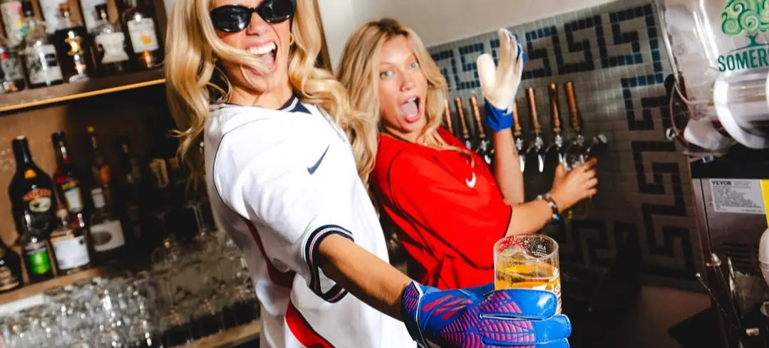 Two people in sports jerseys and gloves serving beer from taps behind a bar, one holding a glass of beer and smiling.