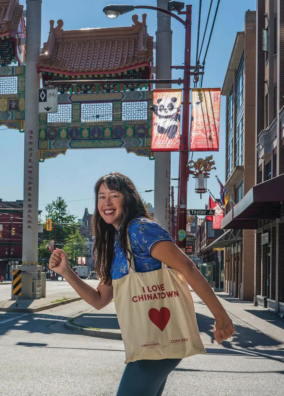 Person smiling and walking near a traditional Chinese archway in a Chinatown district, carrying a tote bag that says I LOVE CHINATOWN.