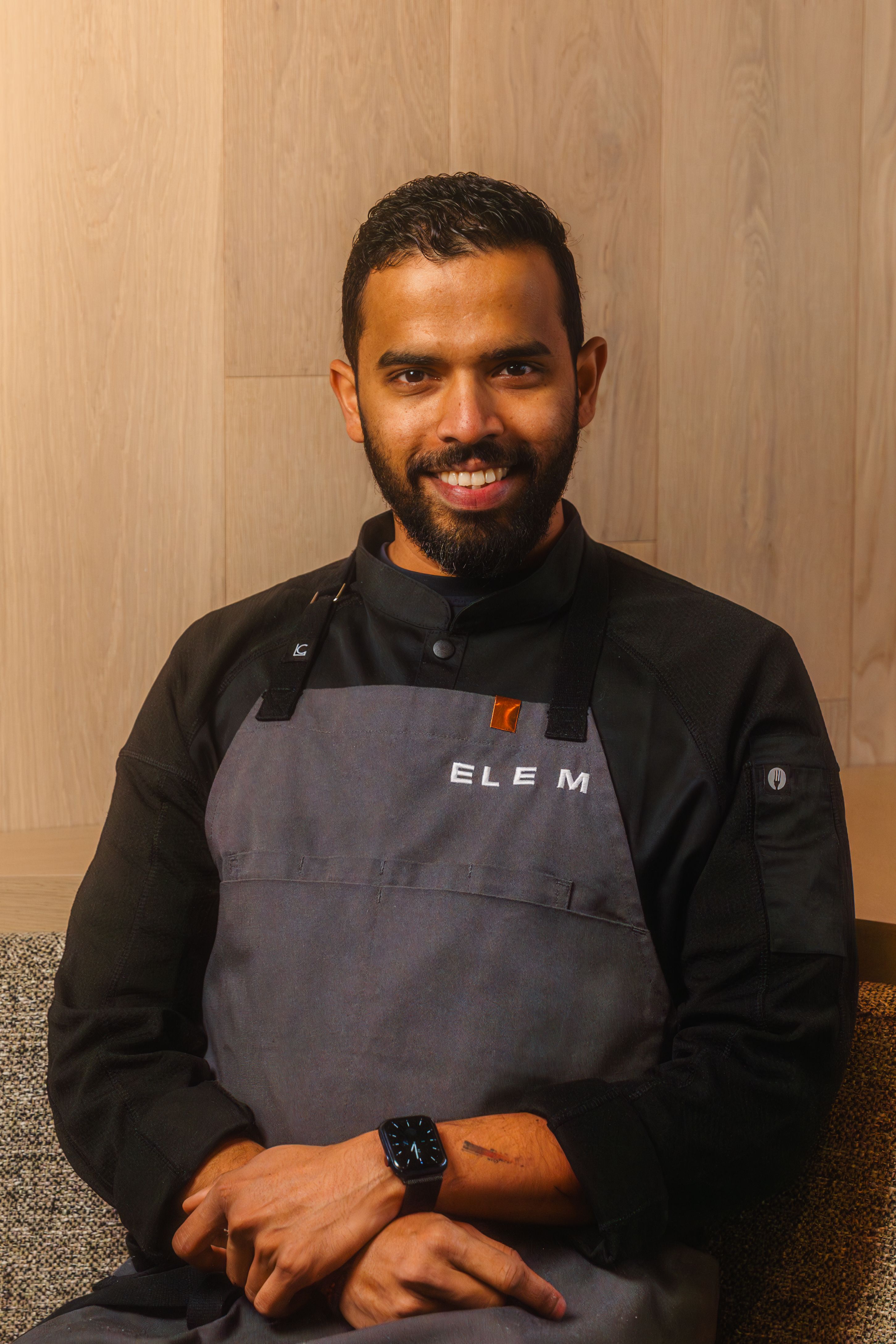 Smiling man with beard wearing a black chef jacket and gray apron with ELEM text, sitting with hands crossed.