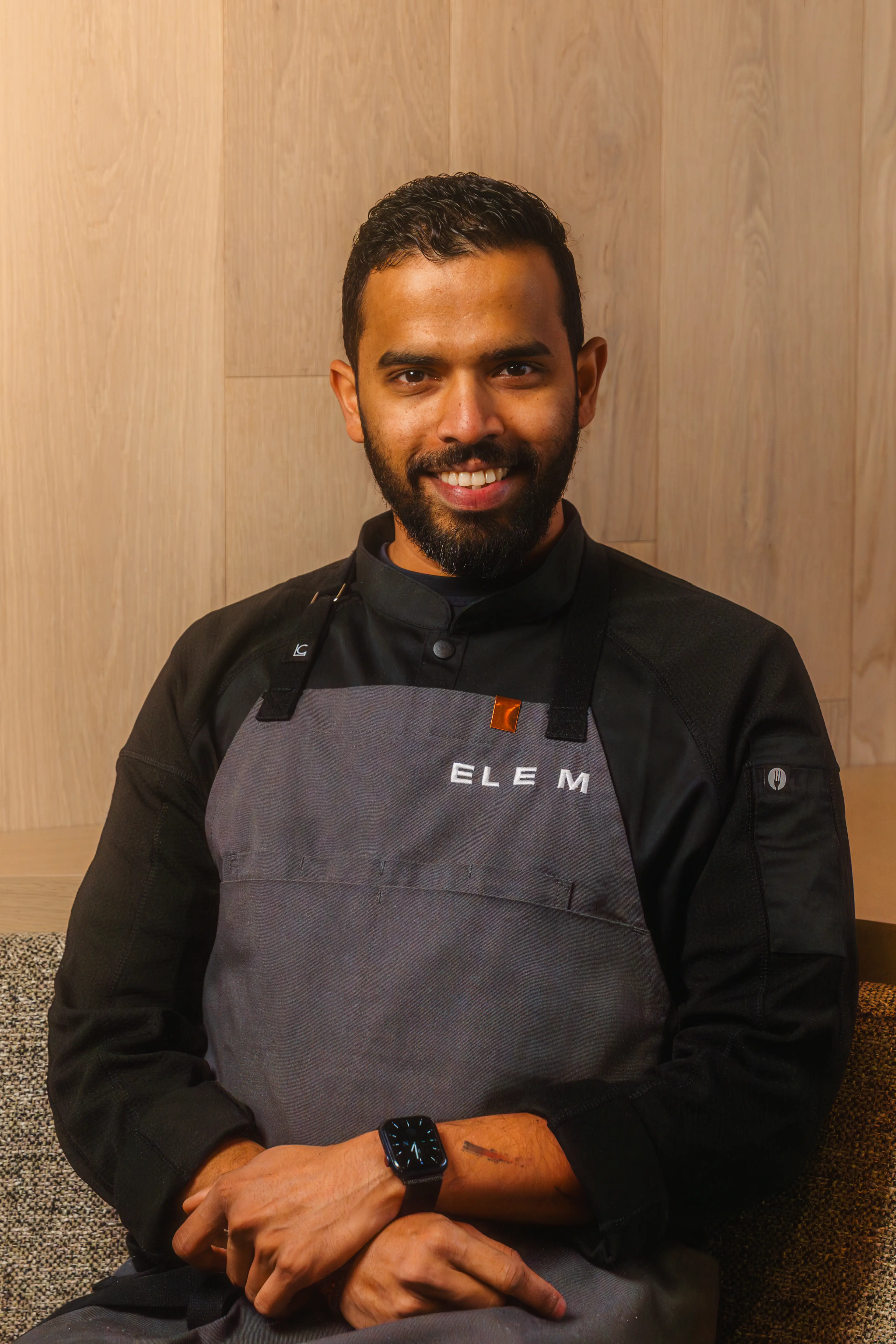 Smiling man with beard wearing a black chef coat and gray apron with ELE M text, sitting with hands crossed.