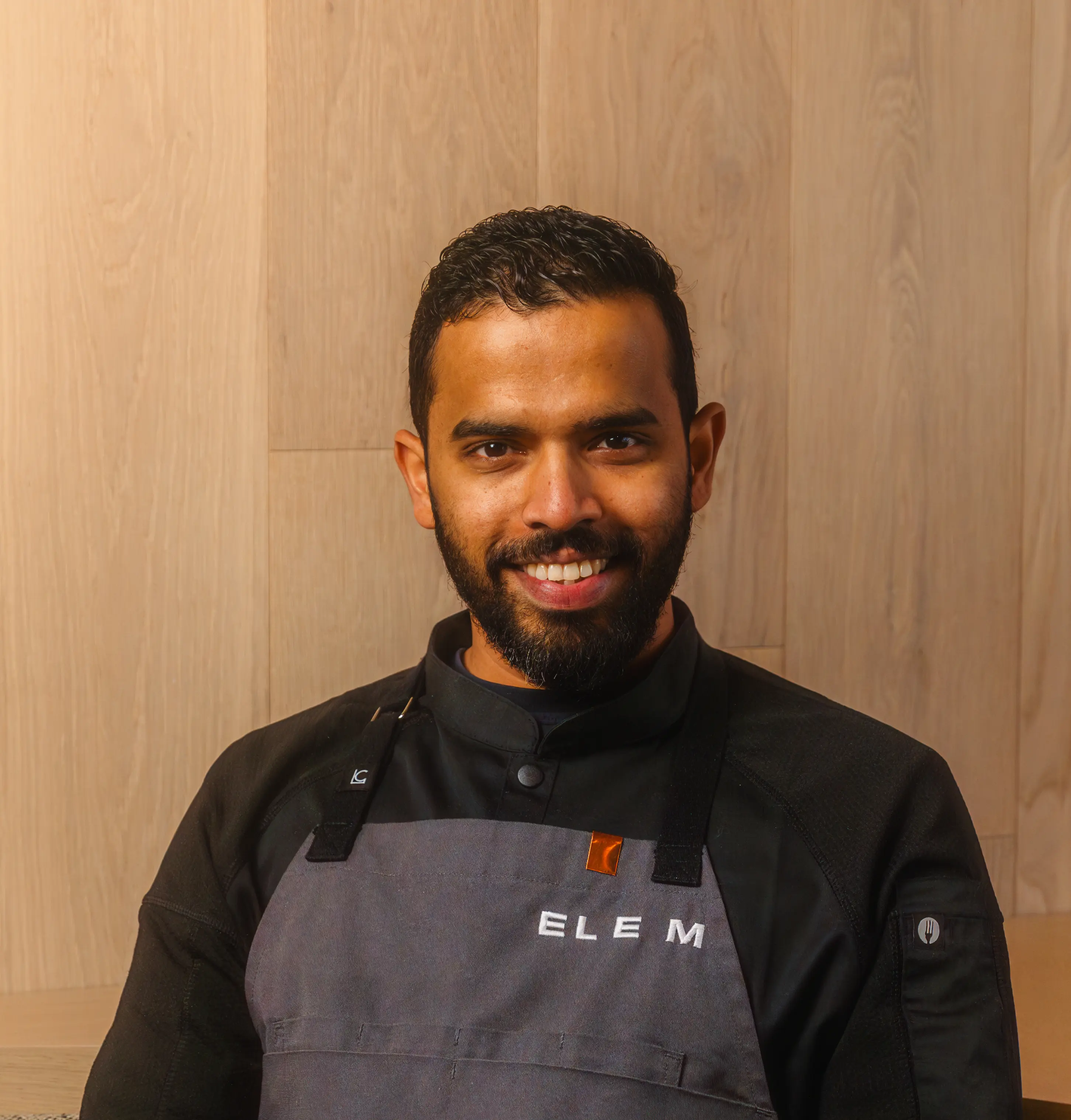 Smiling man with beard wearing a black chef jacket and gray apron with ELEM text, sitting with hands crossed.