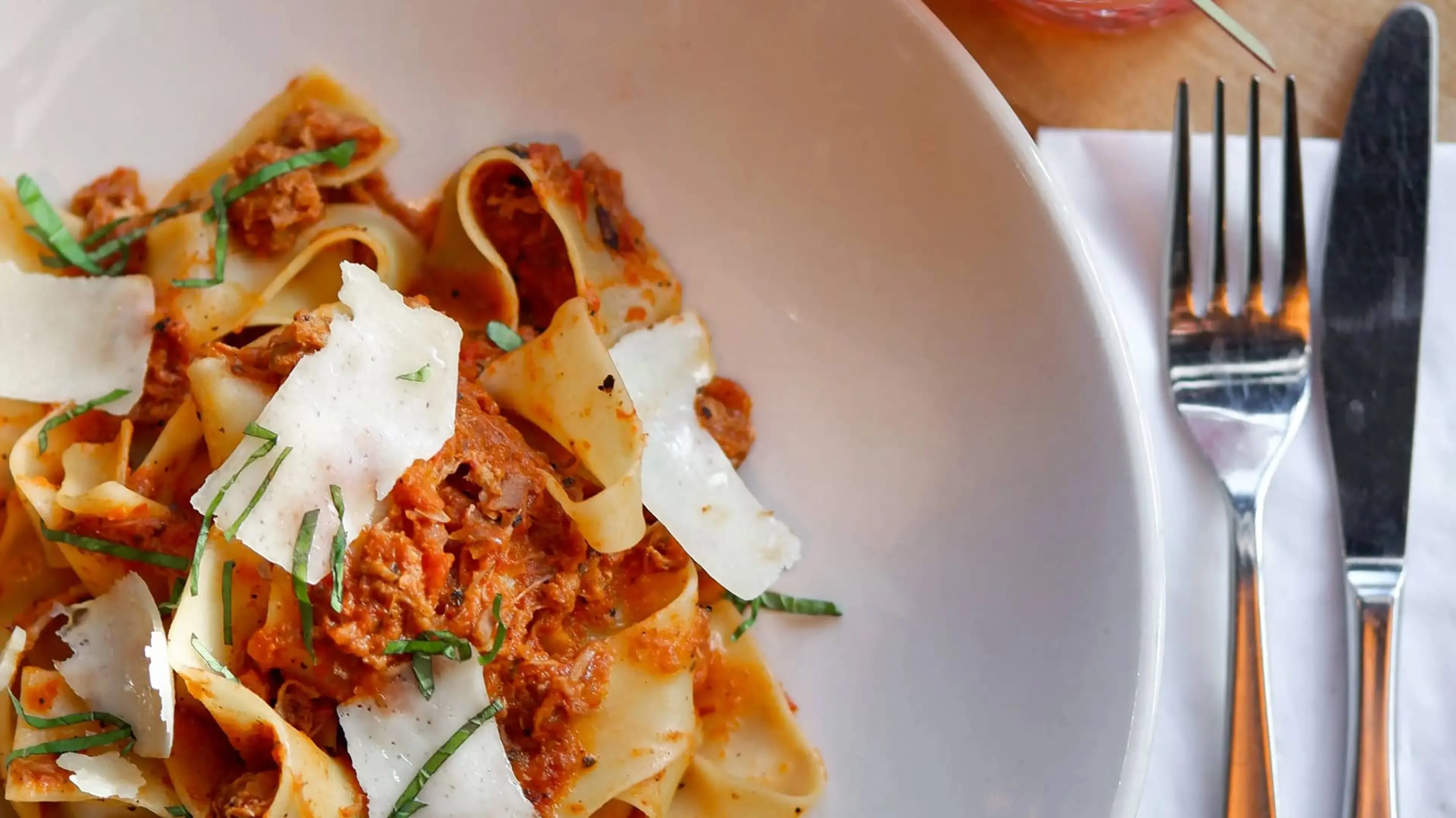Pasta with red sauce, shaved cheese, and basil in a white bowl with fork and knife on a napkin beside it.