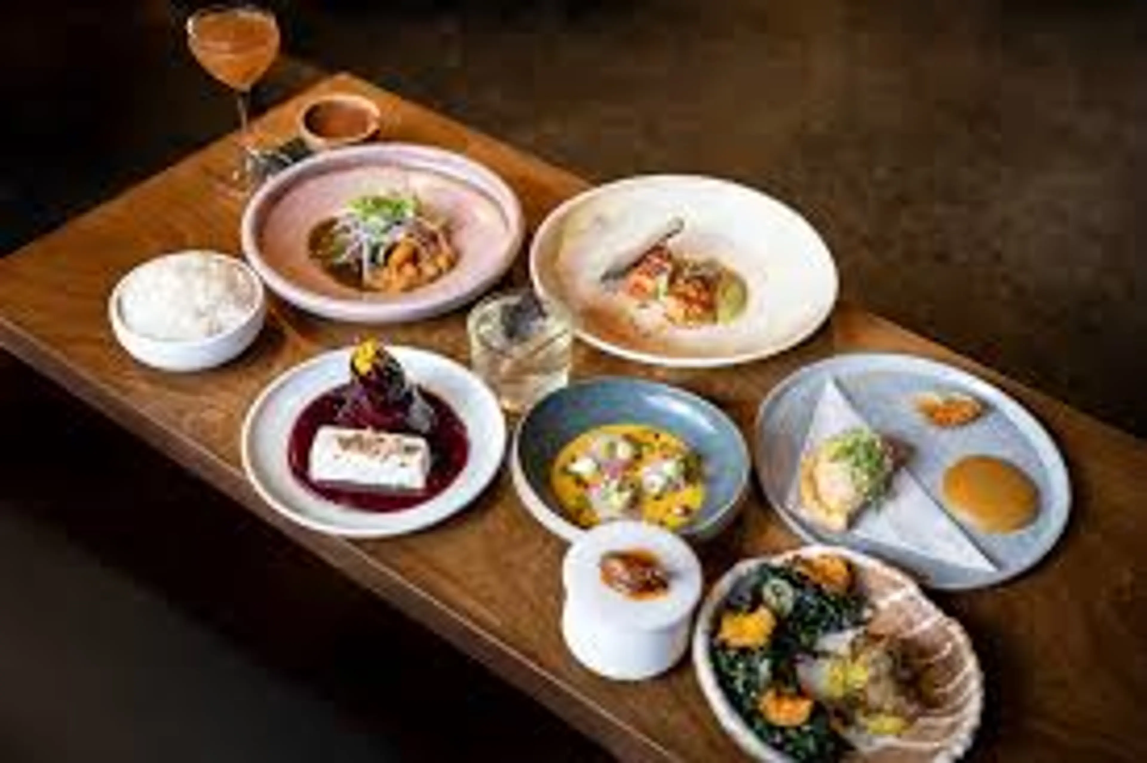 Wooden table with various plated gourmet dishes and a cocktail glass, including rice, seafood, and desserts.