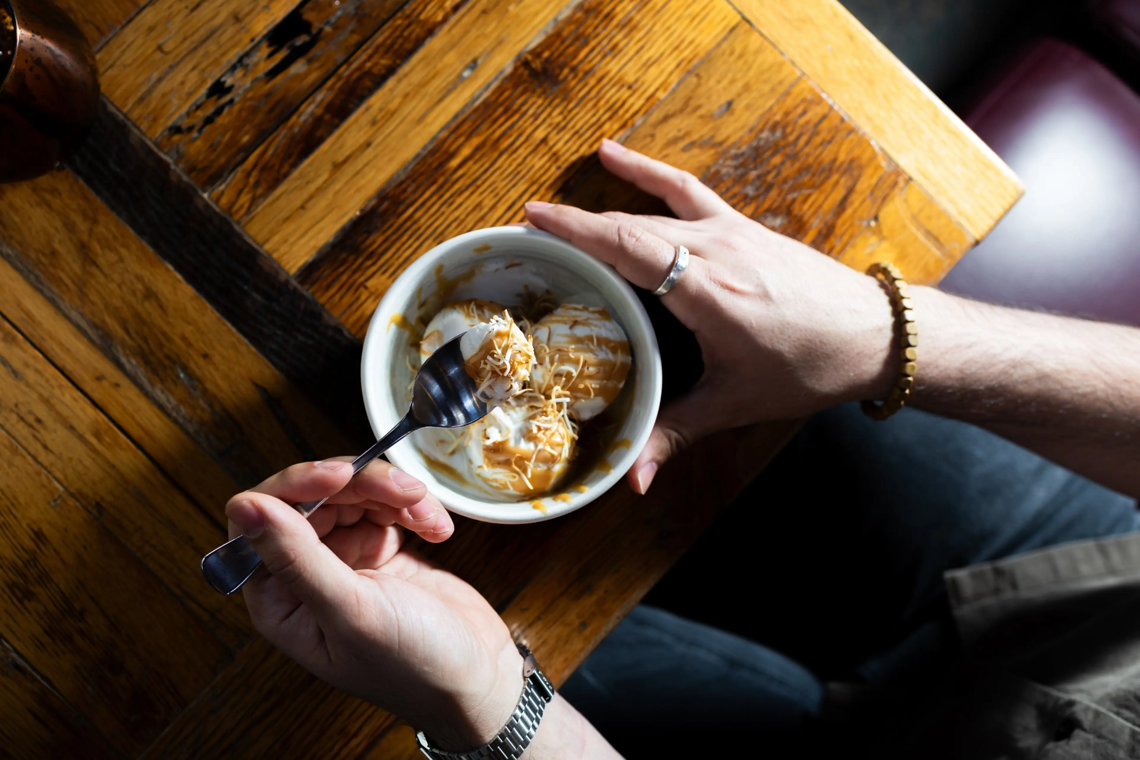 Person holding a bowl of ice cream topped with caramel sauce and shredded coconut, scooping with a spoon over a wooden table.