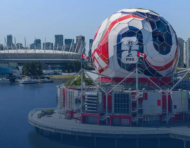 Large spherical building painted like a soccer ball with FIFA logo near waterfront and stadium in background