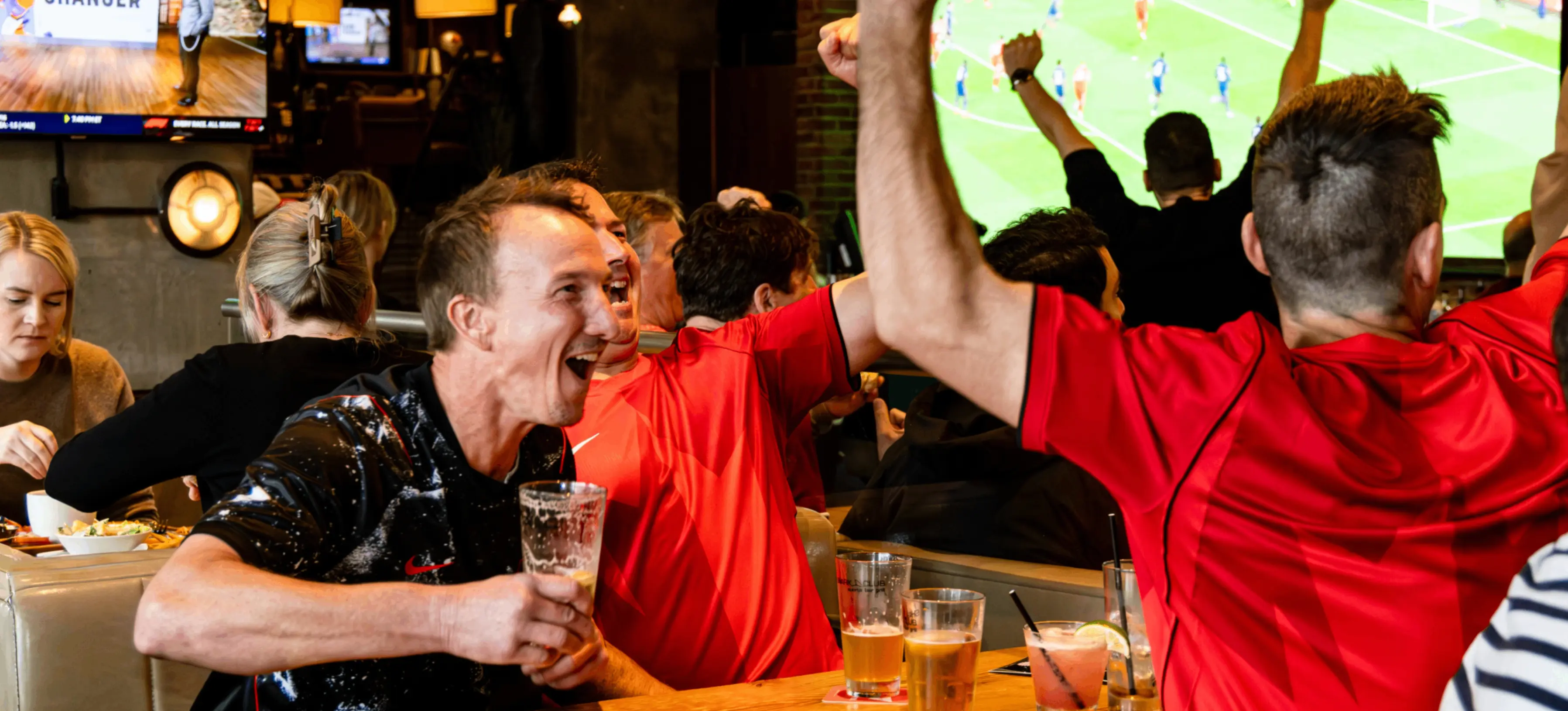 A group of people in a bar cheering and celebrating while watching a soccer game on large TV screens, with drinks on the table.
