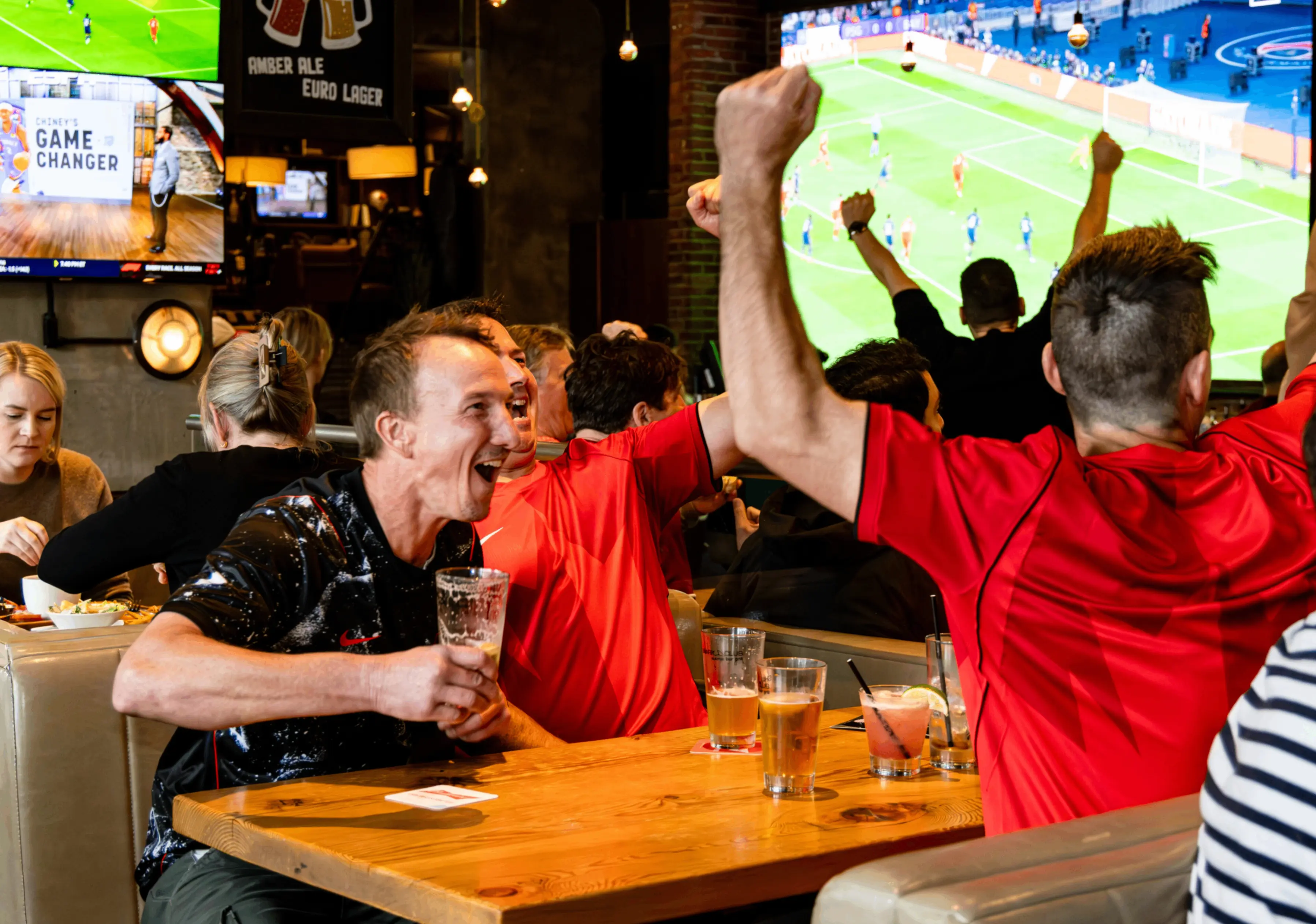 A group of people in a bar cheering and celebrating while watching a soccer game on large TV screens, with drinks on the table.