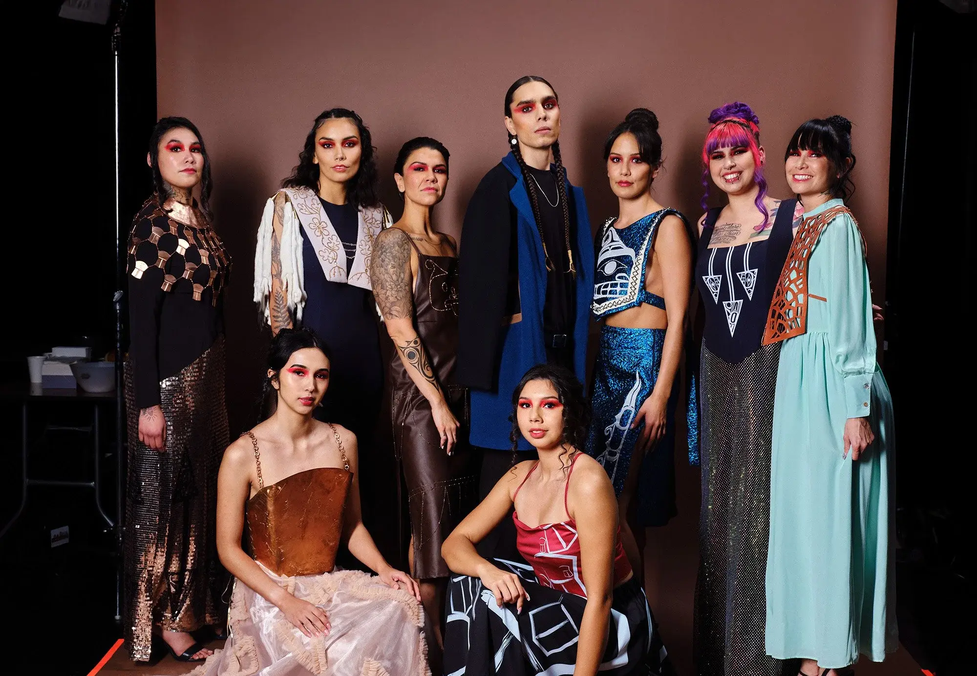 Group of nine people posing in colorful, artistic outfits with bold red eye makeup against a brown backdrop.