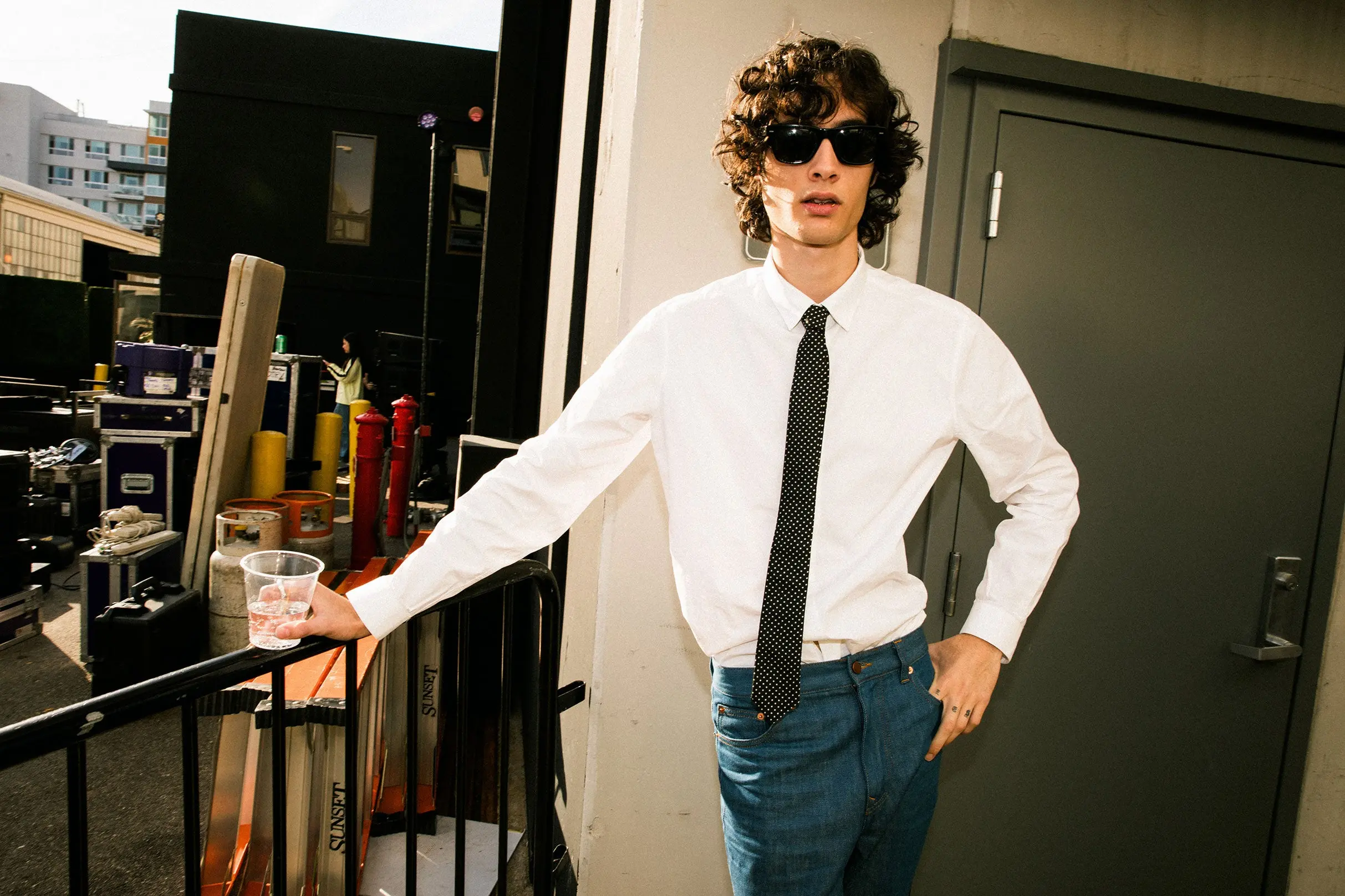 Person with curly hair wearing sunglasses, white shirt, black polka dot tie, and blue jeans, holding a plastic cup and leaning on a railing.