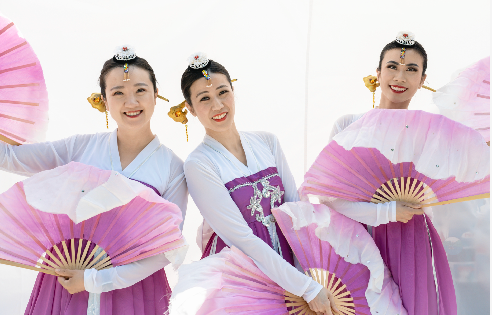Three people in traditional purple and white attire holding pink and white fans, smiling against a white background.