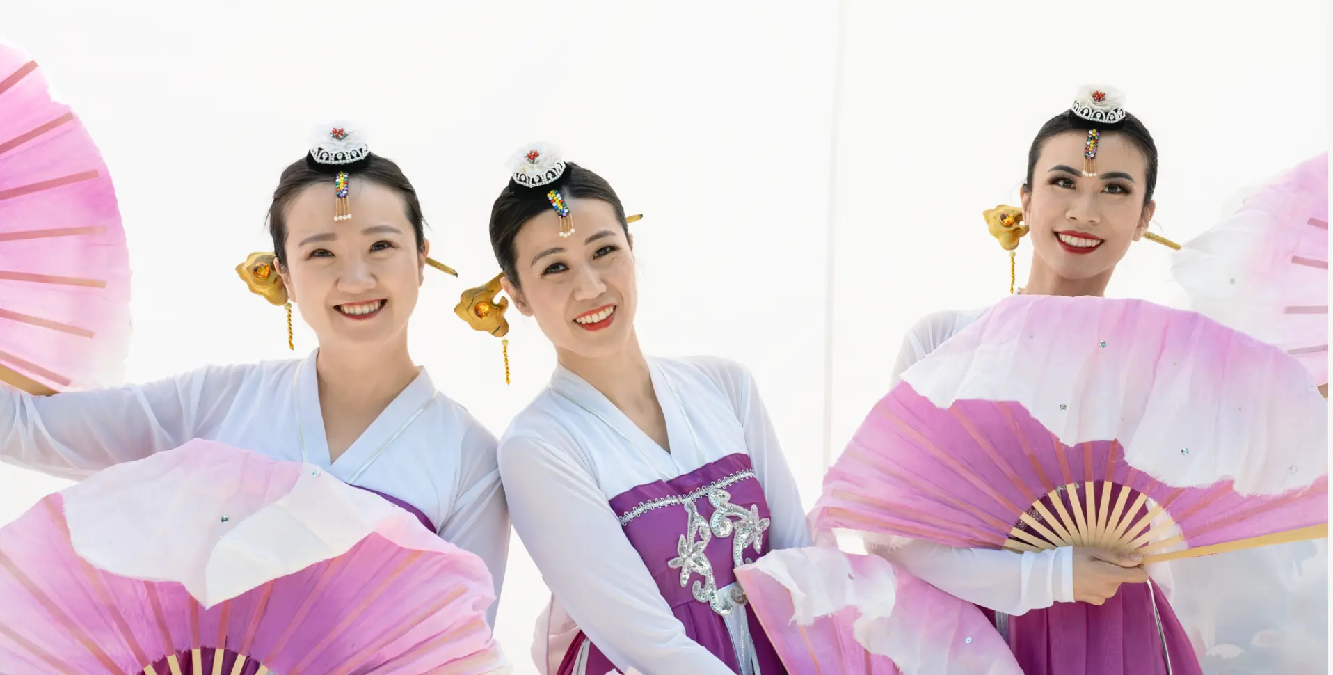 Three people in traditional purple and white attire holding pink and white fans, smiling against a white background.
