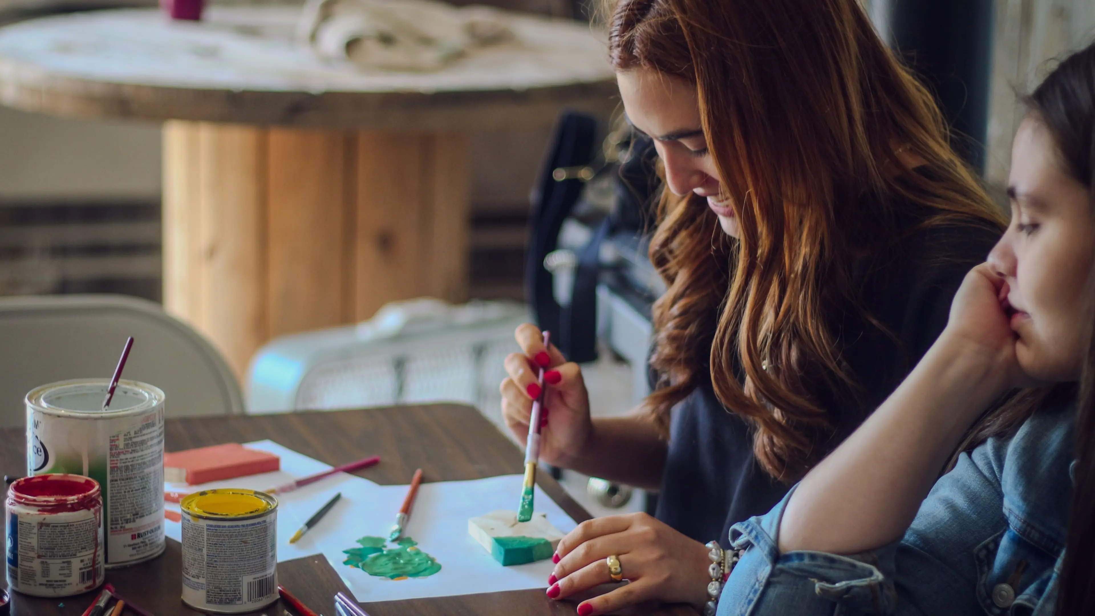 Two people sitting at a table with paint cans and brushes, one painting a small object with green paint.