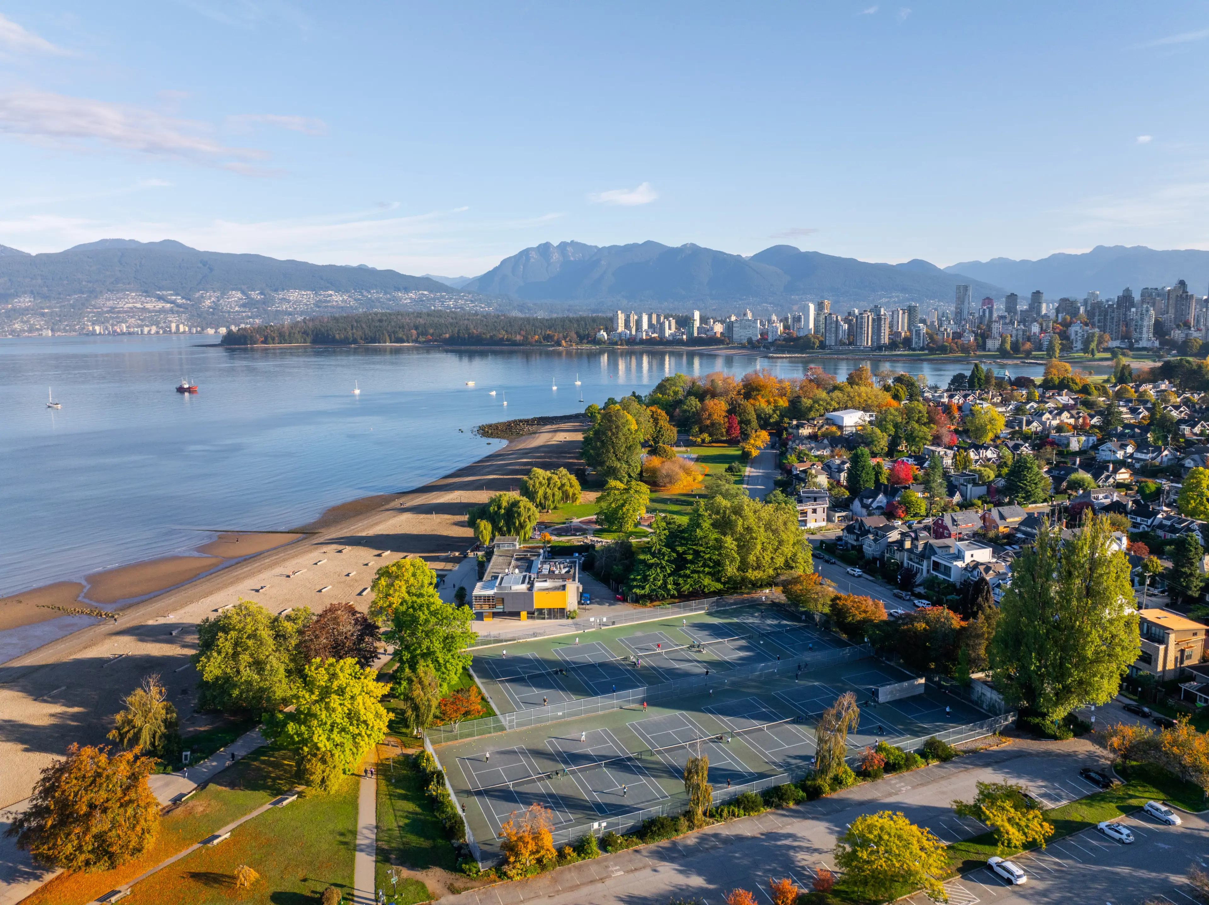 Aerial view of a coastal city with tennis courts, a sandy beach, trees with autumn foliage, and mountains in the background.