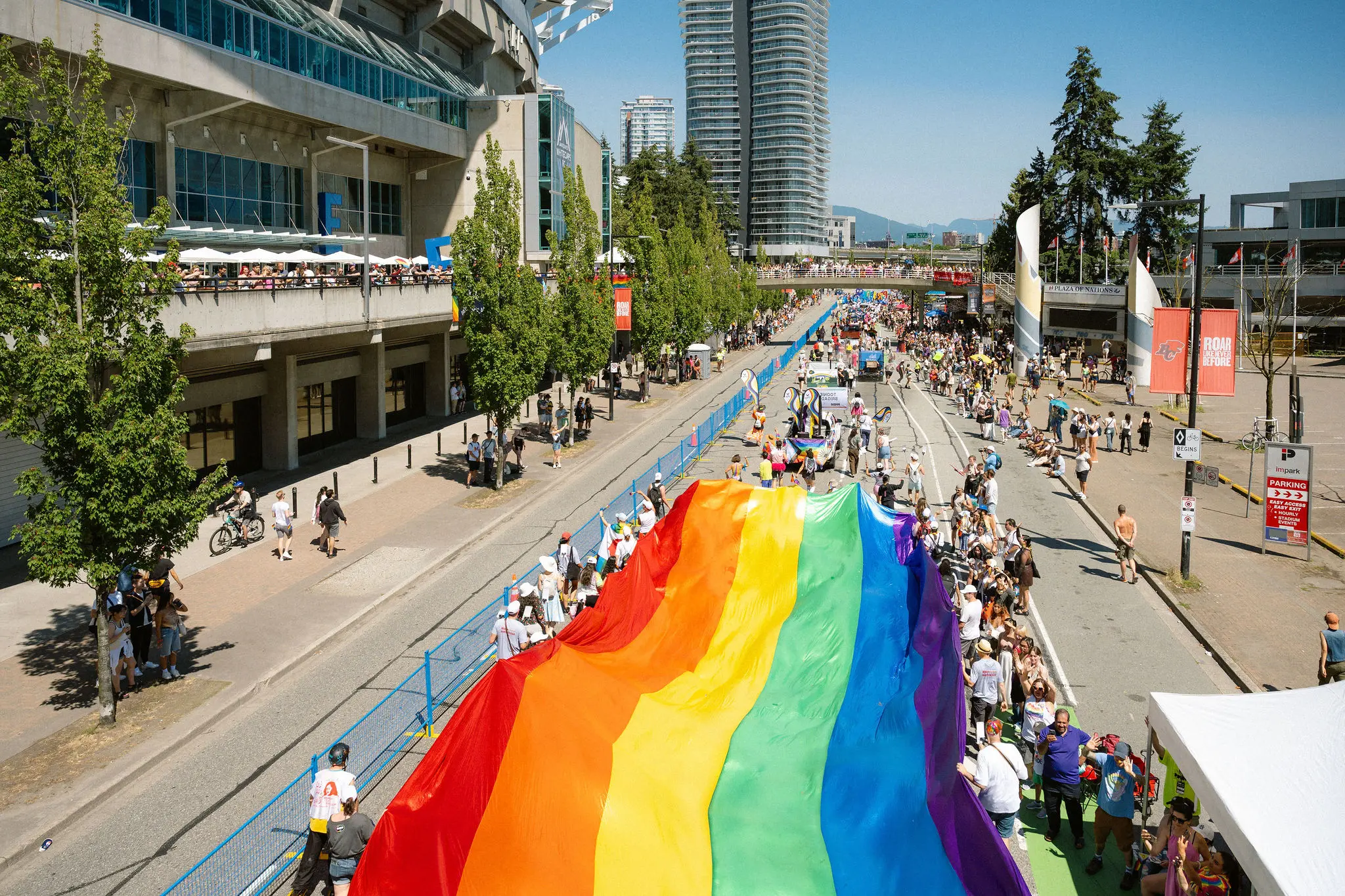 Large rainbow flag held by a crowd during a street parade with buildings and trees on the sides.
