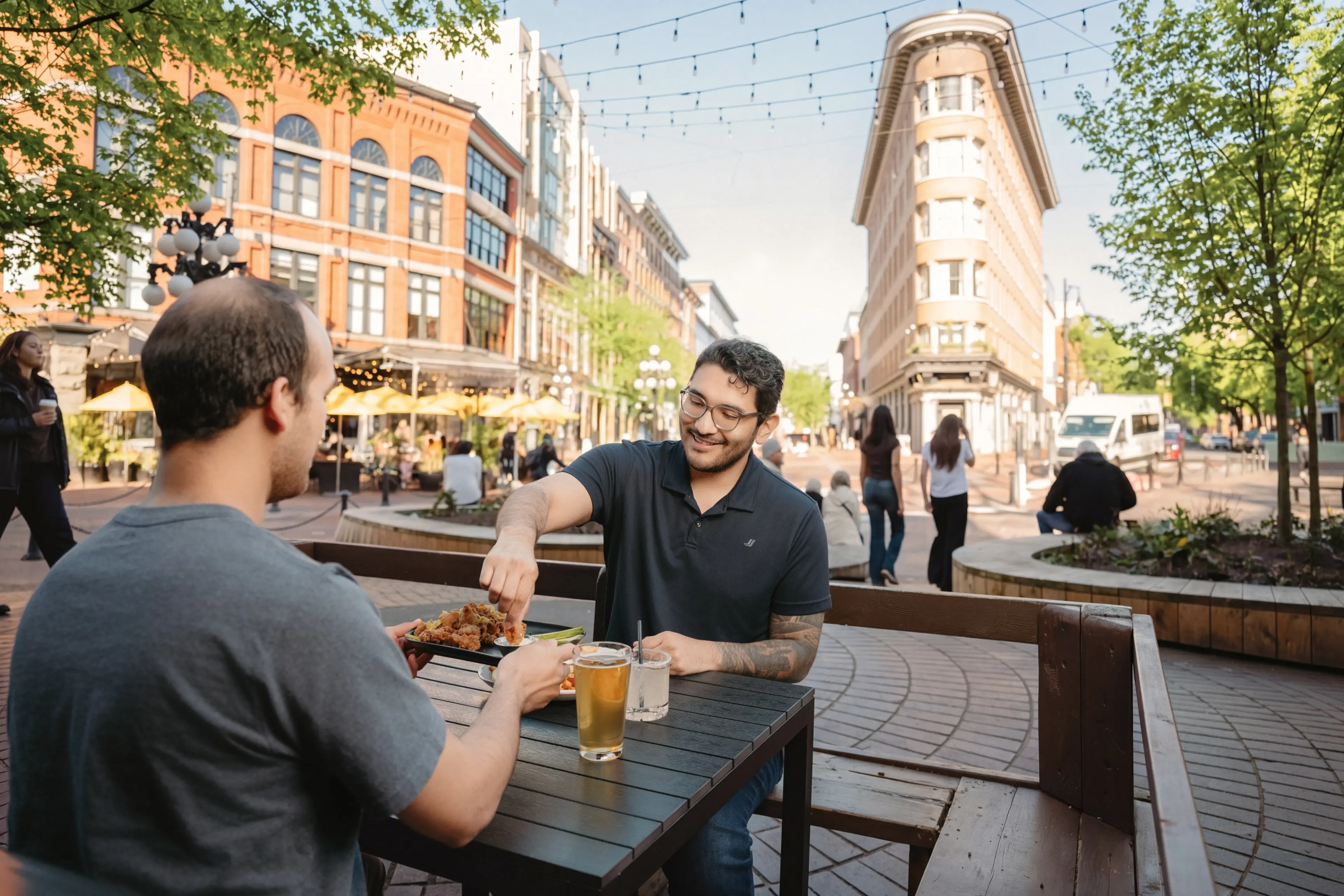 Two men sitting on a patio at the Six Acres restaurant in Gastown, Vancouver sharing food and drinks on a sunny day.