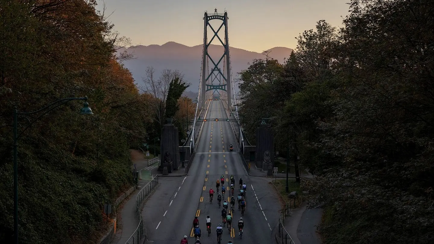 Group of cyclists riding on a bridge surrounded by trees with mountains in the background at dusk.